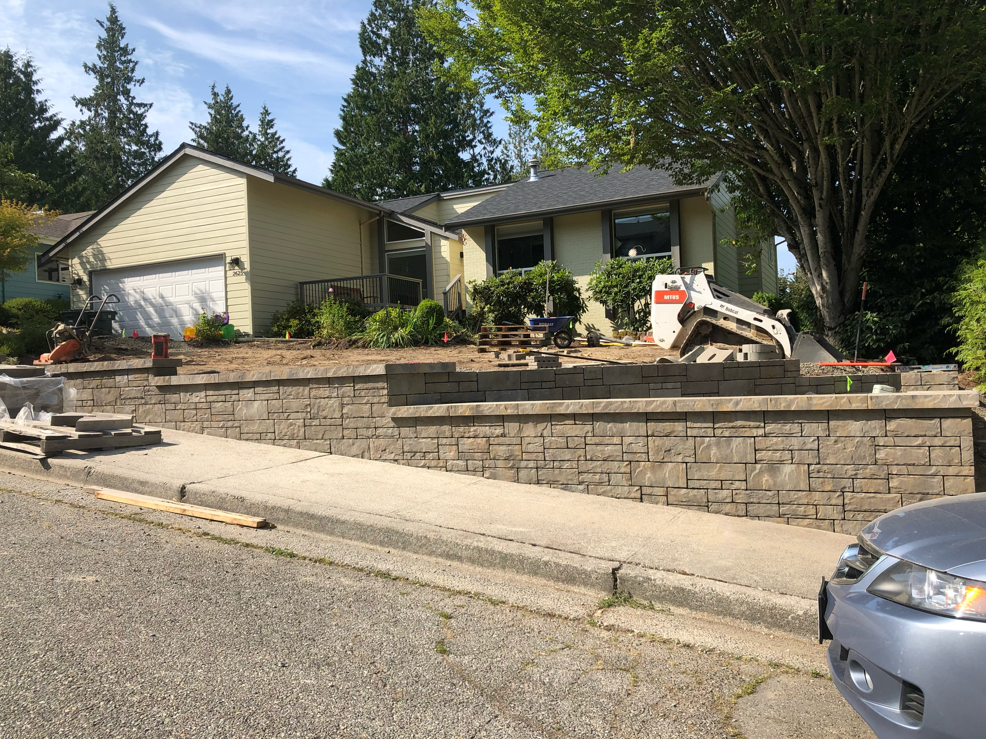 Residential property with stone retaining wall; construction equipment visible. Sunny day; work by West Coast Landscape & Hardscape. 