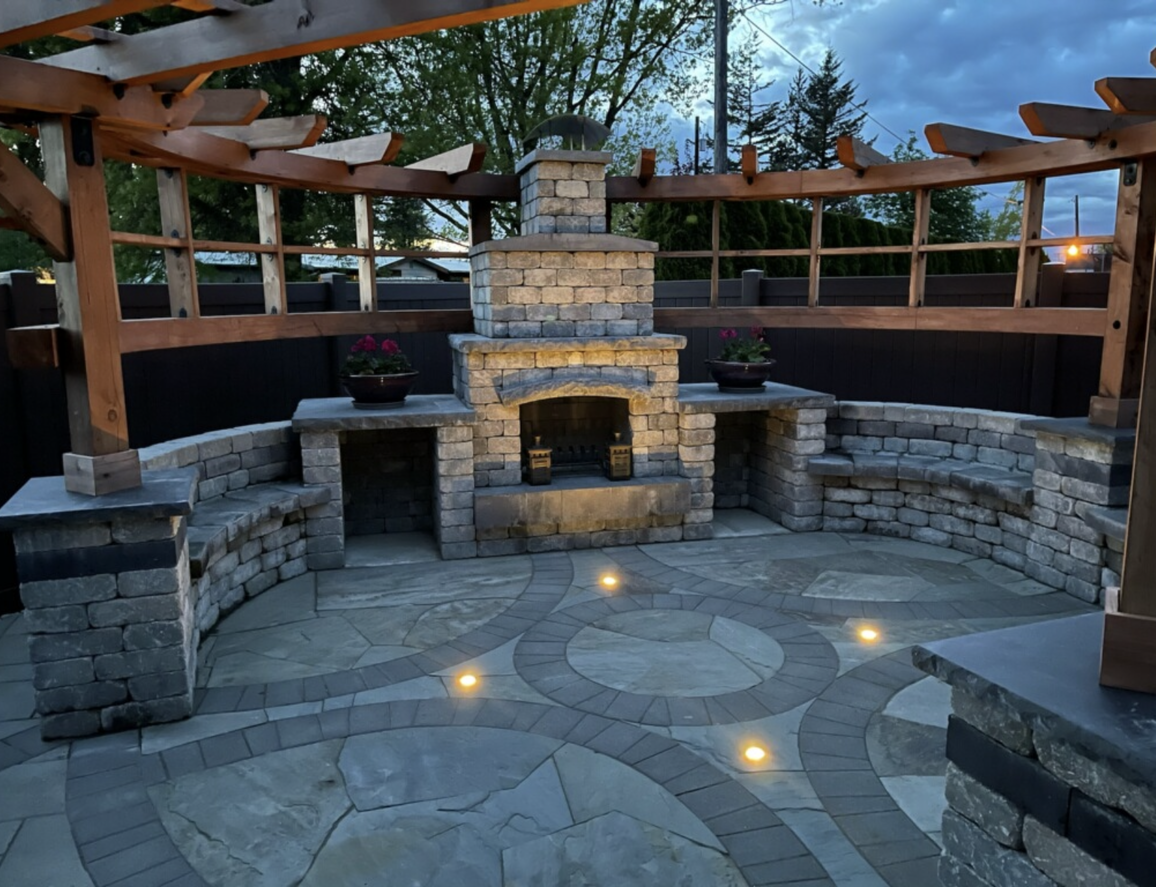 Outdoor stone fireplace and seating area under a wooden pergola, illuminated by spotlights; work by West Coast Landscape & Hardscape. 