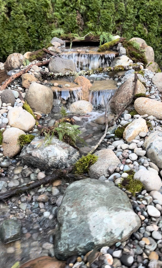 Water cascades down a multi-tiered rocky waterfall. Green moss and pebbles surround it; by West Coast Landscape & Hardscape. 