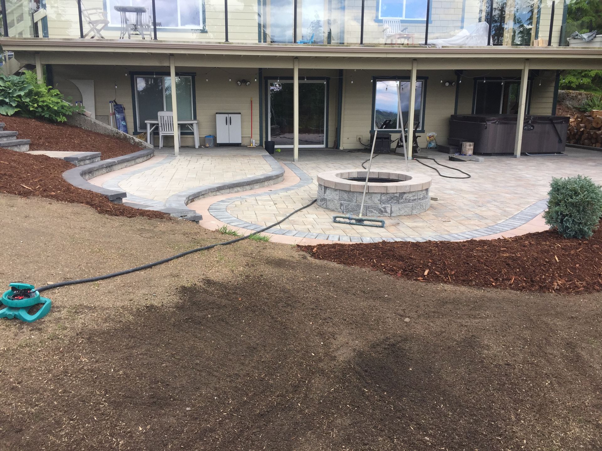 Patio with fire pit, stone paving, and house in background. Mulch and sprinkler in foreground; work by West Coast Landscape & Hardscape. 