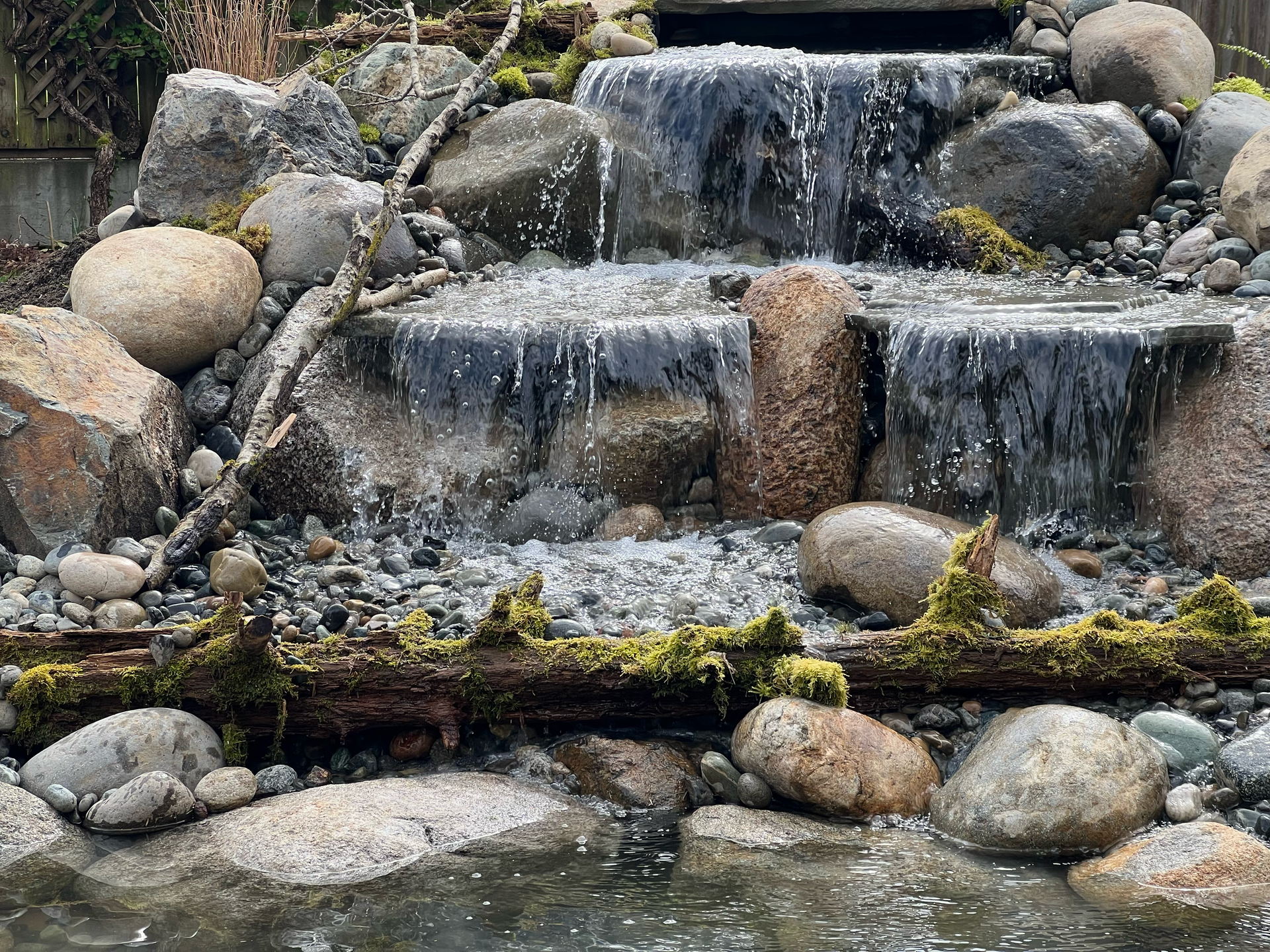 A multi-tiered waterfall cascading over rocks into a pond; moss and wood accents; work by West Coast Landscape & Hardscape. 