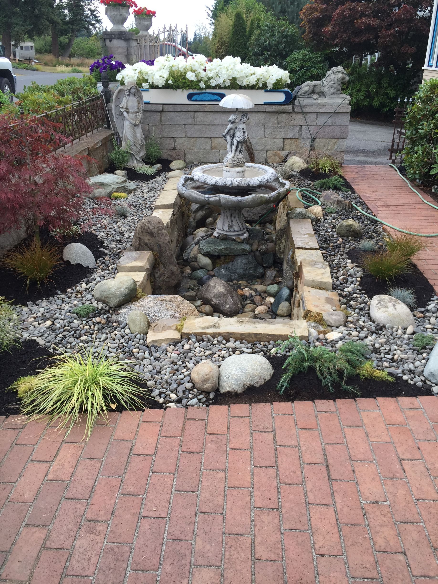 Stone fountain in a brick-lined garden bed with surrounding plants and a decorative wall; work by West Coast Landscape & Hardscape. 