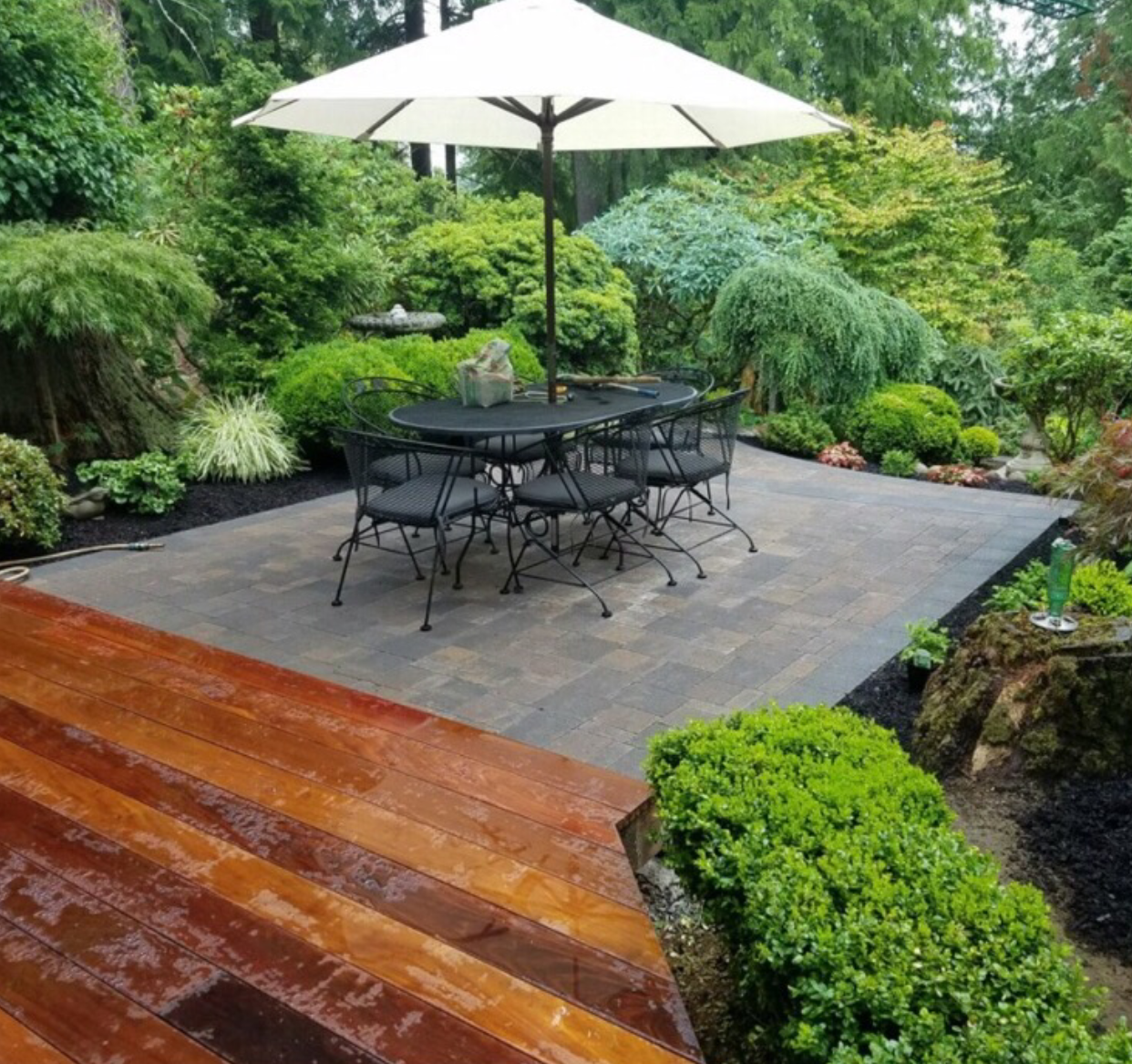 Patio with table, chairs, and umbrella surrounded by lush greenery and a wooden deck; work by West Coast Landscape & Hardscape. 