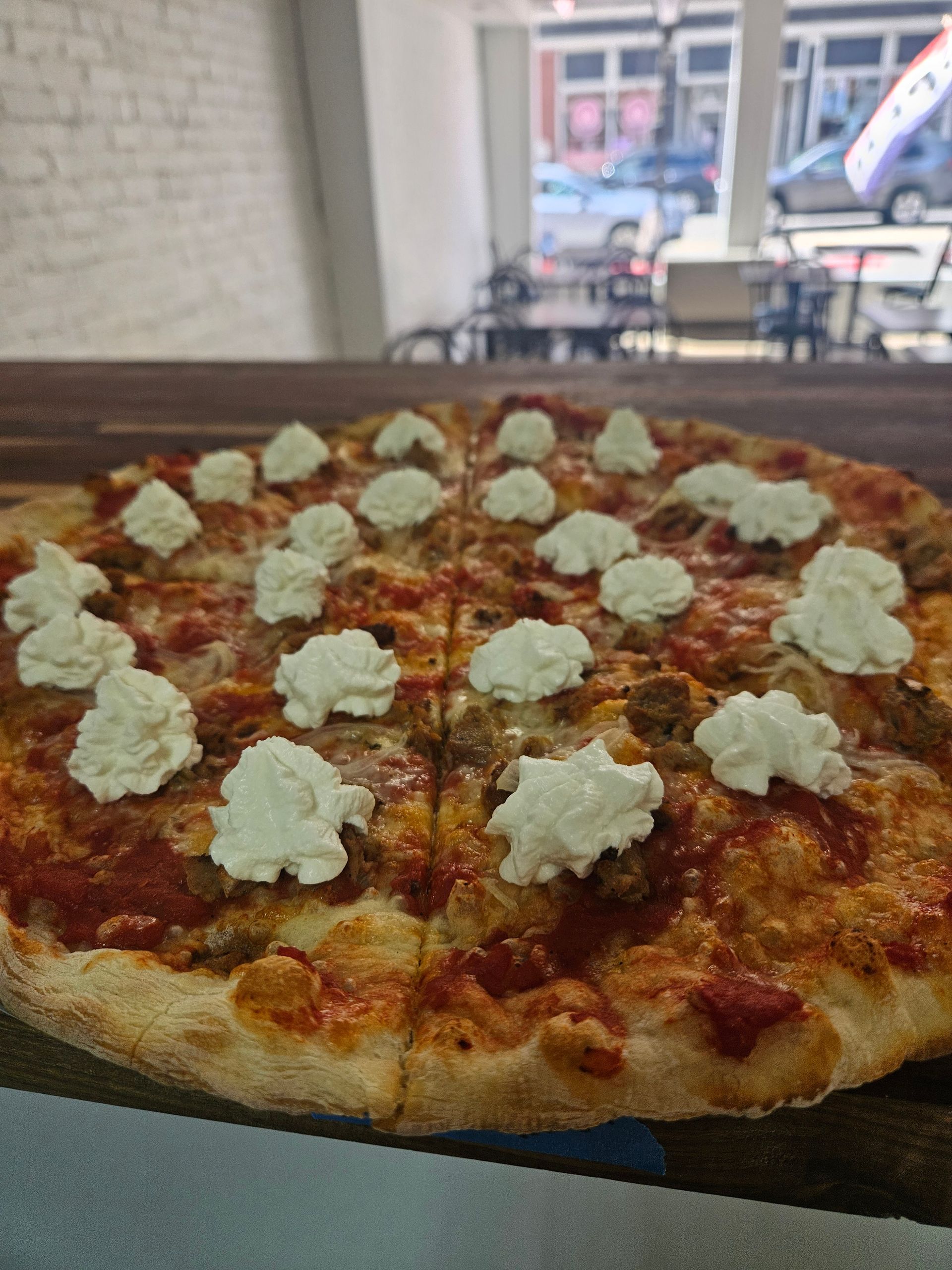 Pizza with white dollops of cheese, on a wood surface near an outdoor view.