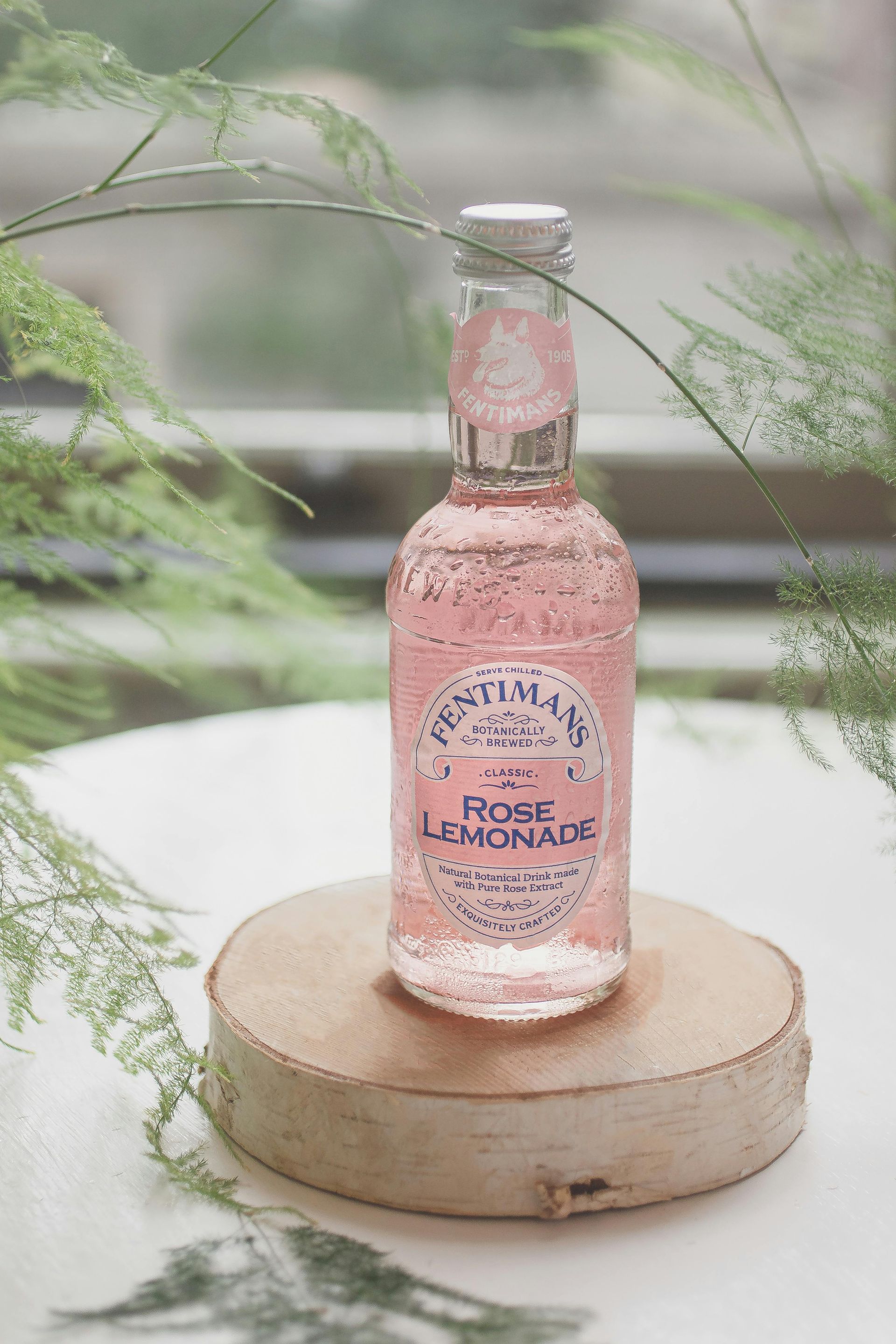 Pink rose lemonade bottle on a wooden stand, surrounded by green ferns.