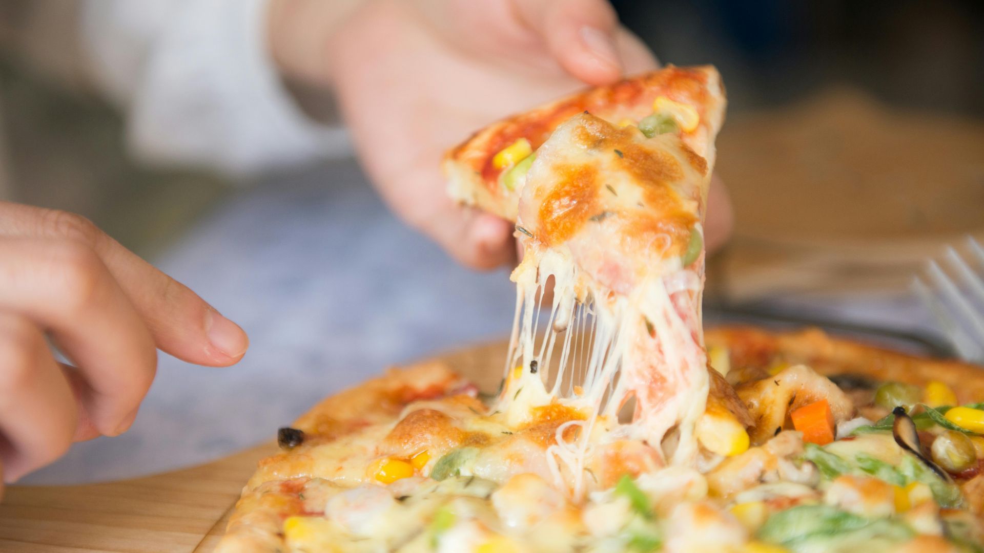 Hand holding a pizza slice, with cheese stretching. Pizza has vegetables.