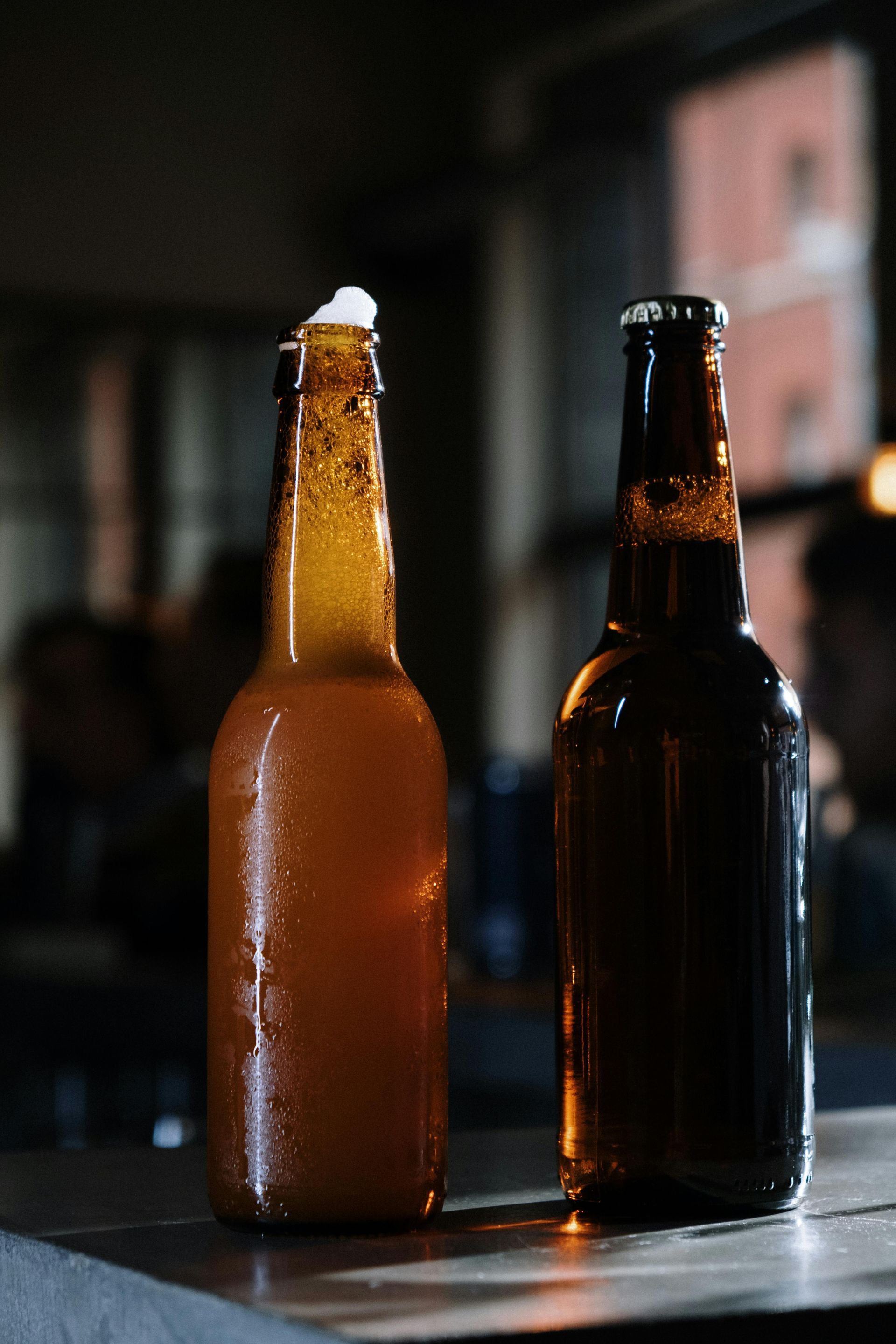 Two beer bottles, one amber, one dark, on a counter, indoors.