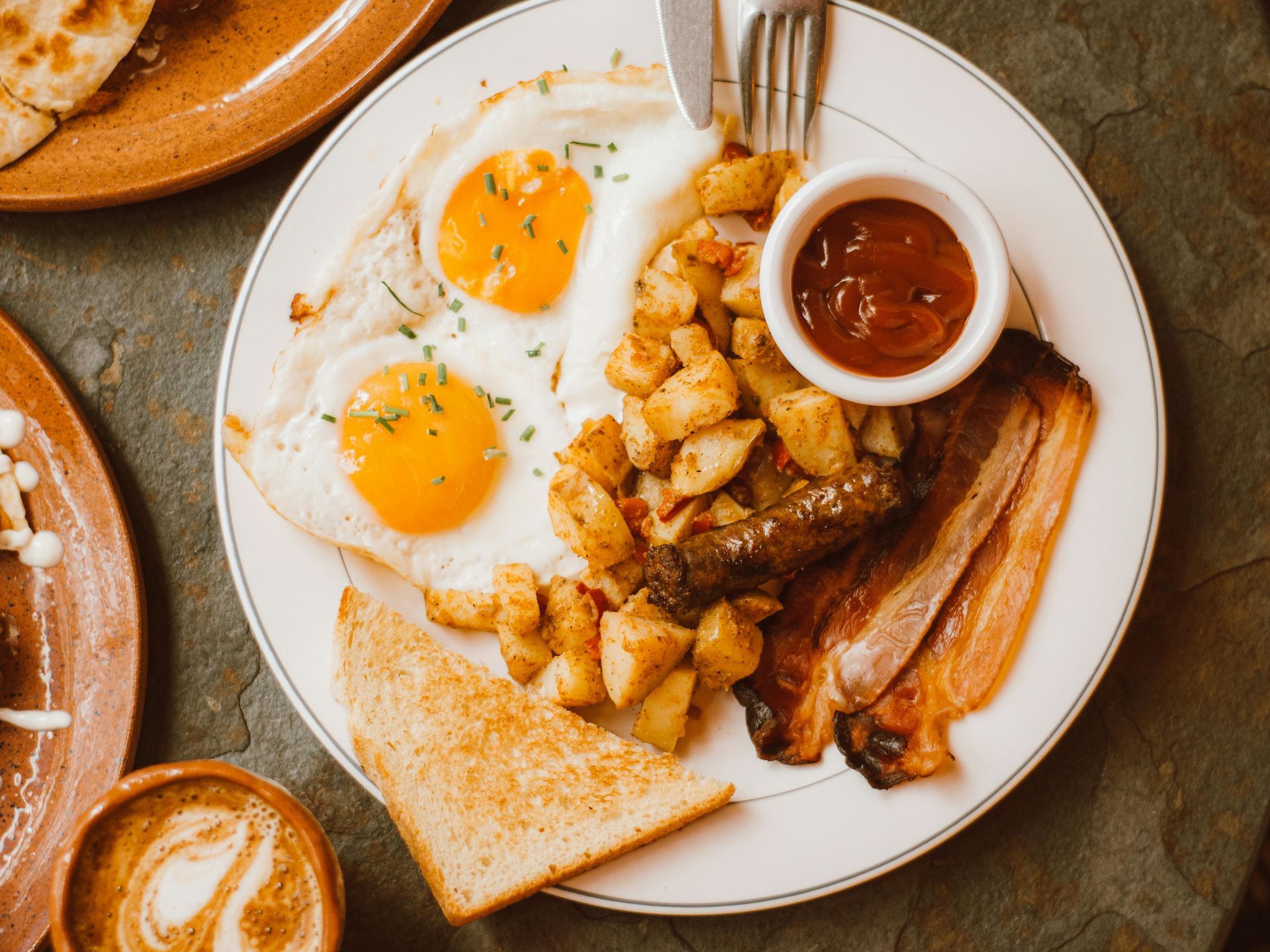 Breakfast plate with eggs, potatoes, bacon, sausage, toast, and ketchup.