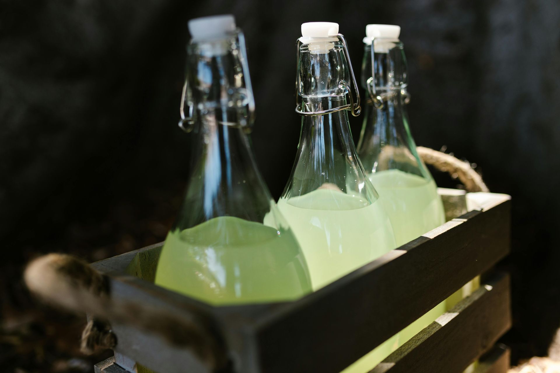 Three glass bottles of light green liquid in a wooden crate with rope handles.