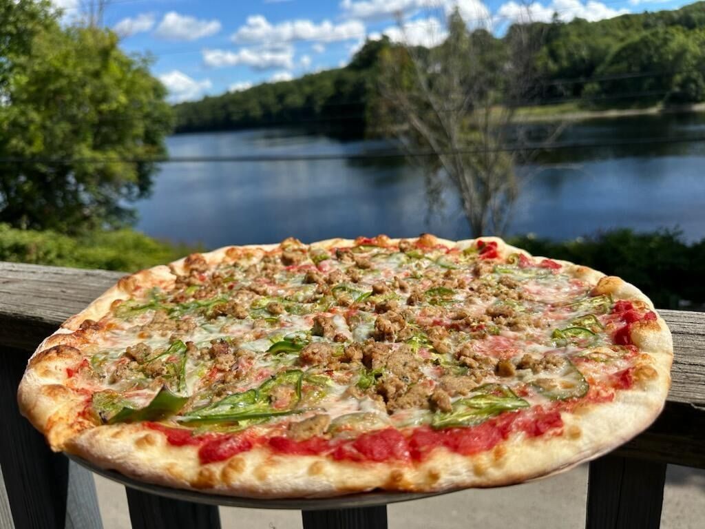 Pizza with toppings on a wooden deck overlooking a lake and trees under a blue sky.