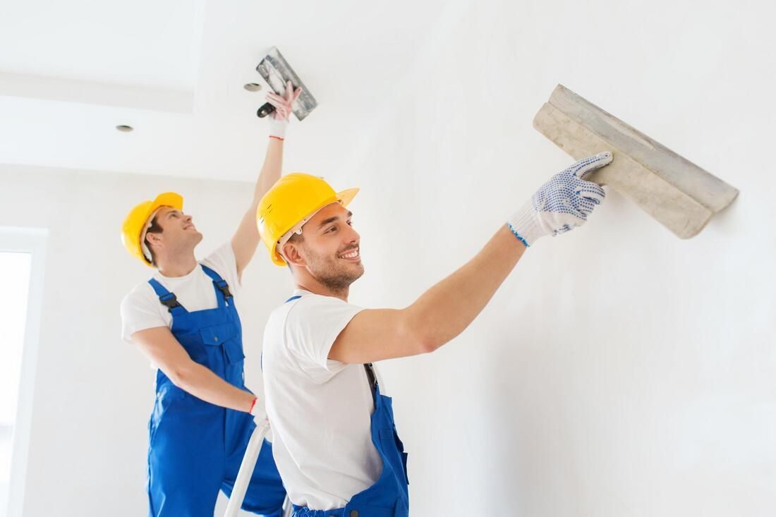 Two workers in blue overalls and yellow hard hats smoothing white plaster on a wall.