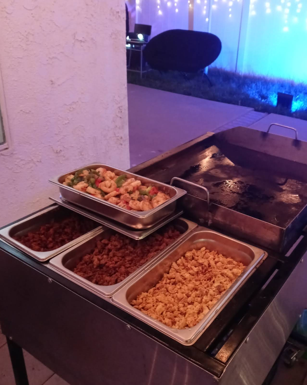 Food on a catering cart: various meats and shrimp, and a flat grill ready for cooking, outdoors.