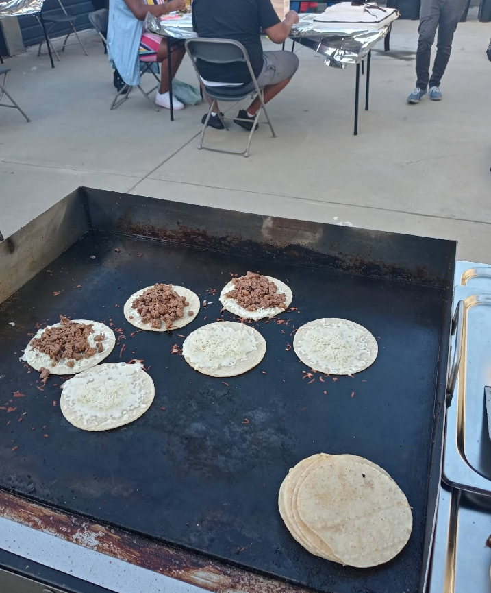 Tacos being cooked on a griddle outdoors, people sitting at tables in the background.