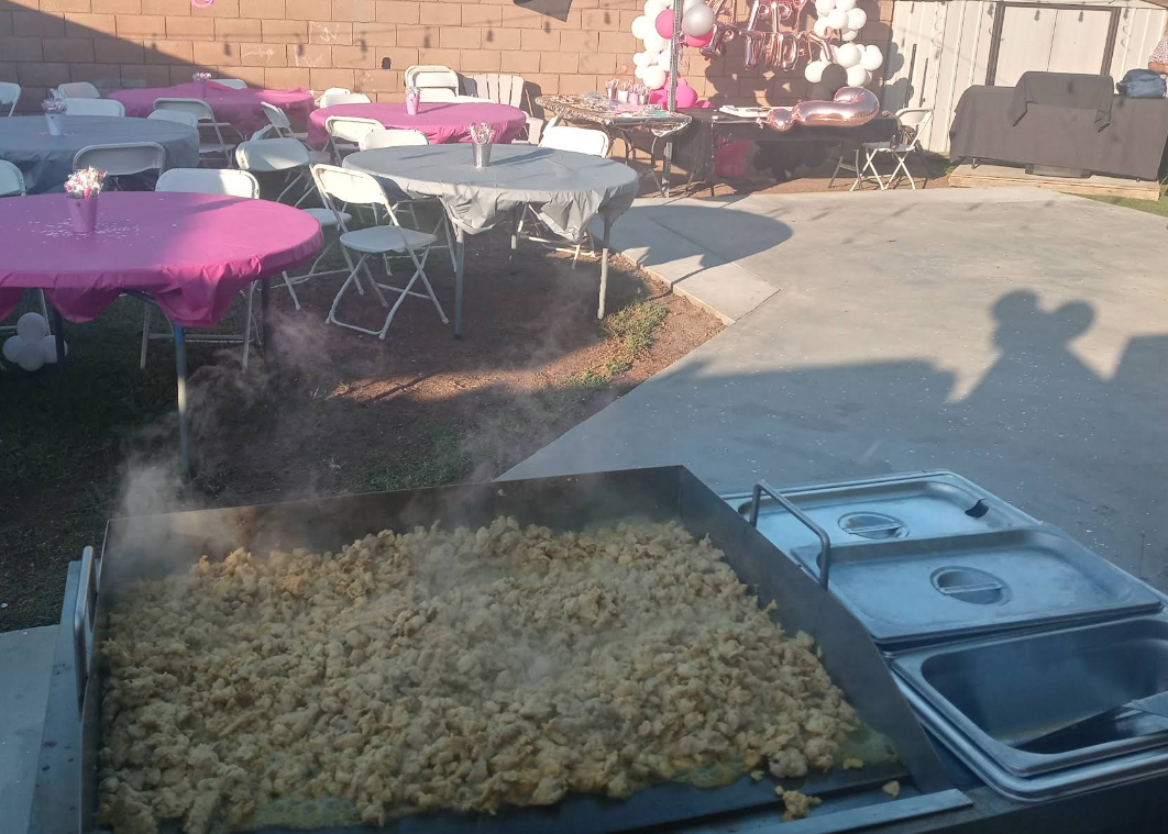 Large outdoor party: food being cooked on a griddle. Tables with pink cloths are set up nearby.