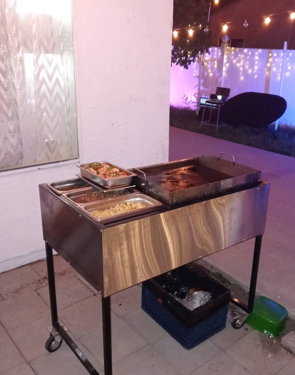 Food cart with griddle and containers of food, outdoor setting with lights.