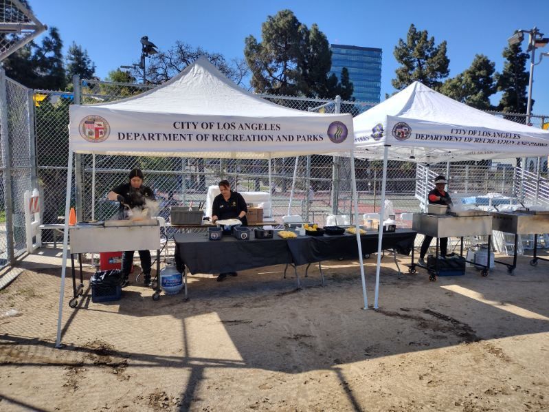 City of Los Angeles Department of Recreation and Parks tents with people working, set up outdoors near a chain-link fence.