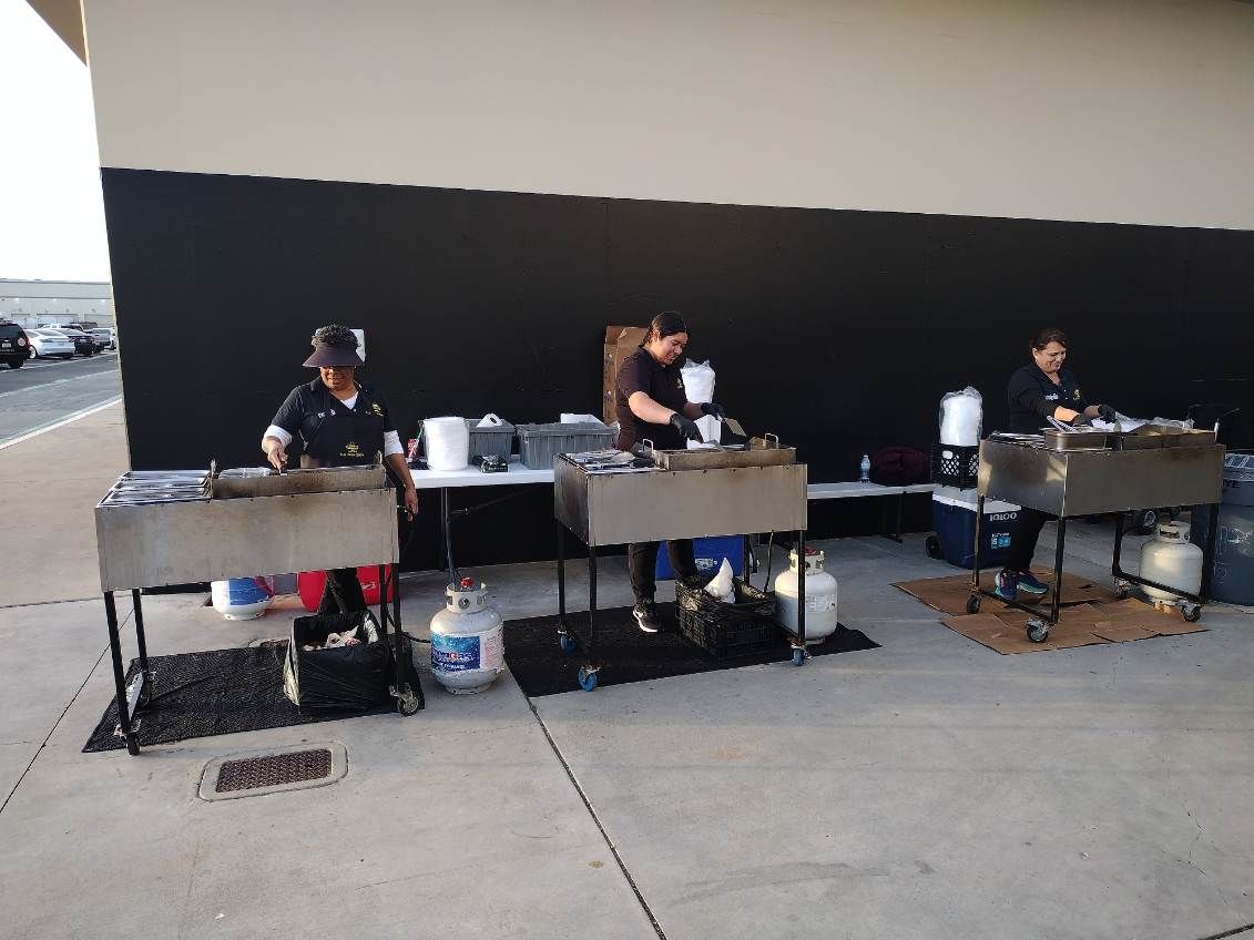 Three people cook food at outdoor food stations. Black wall backdrop.