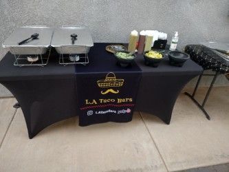 Taco bar setup with chafing dishes, toppings, and branding on a black table.