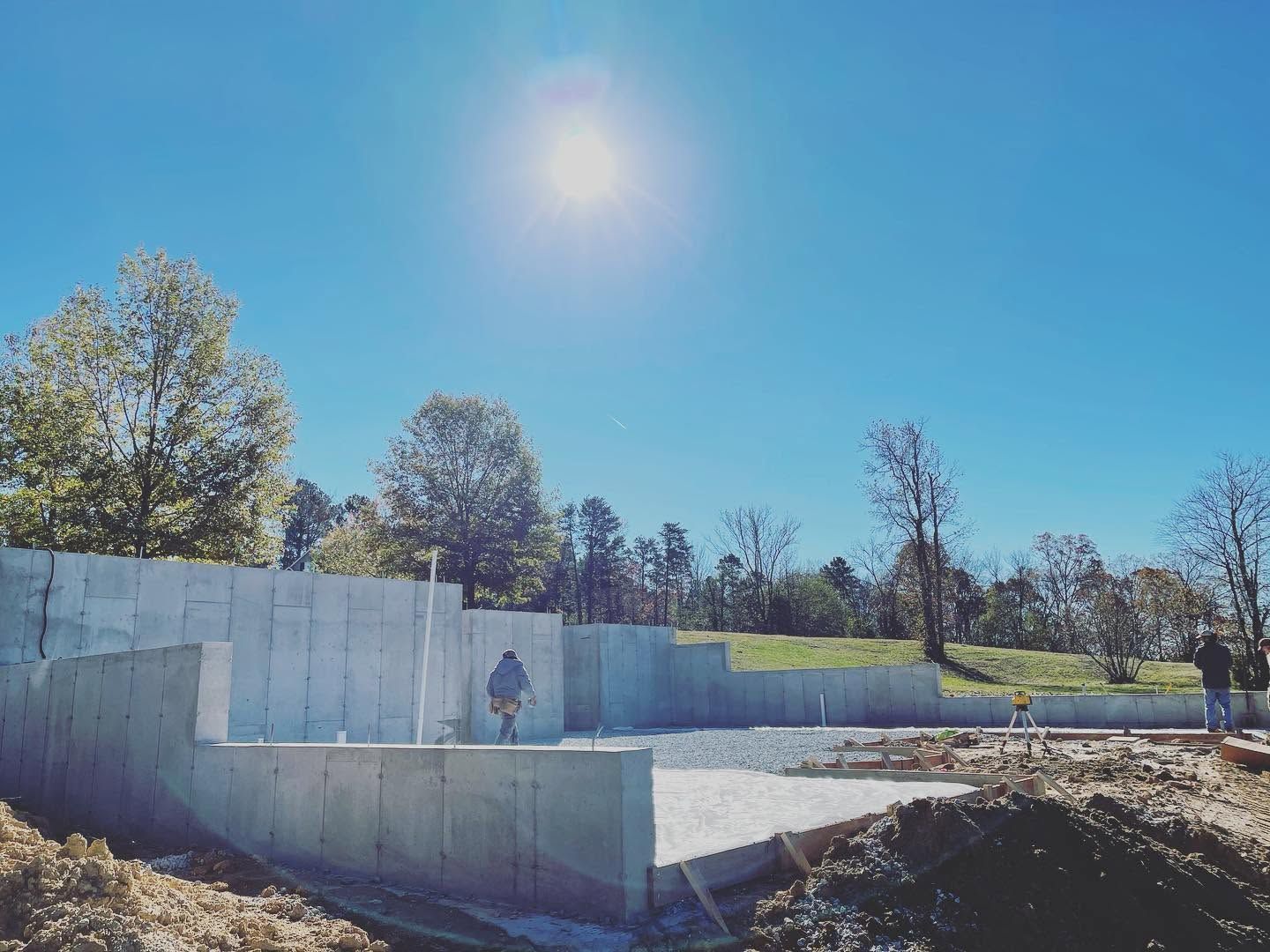 Outdoor concrete skatepark bowl under a bright blue sky, with trees and a person standing on the wall.