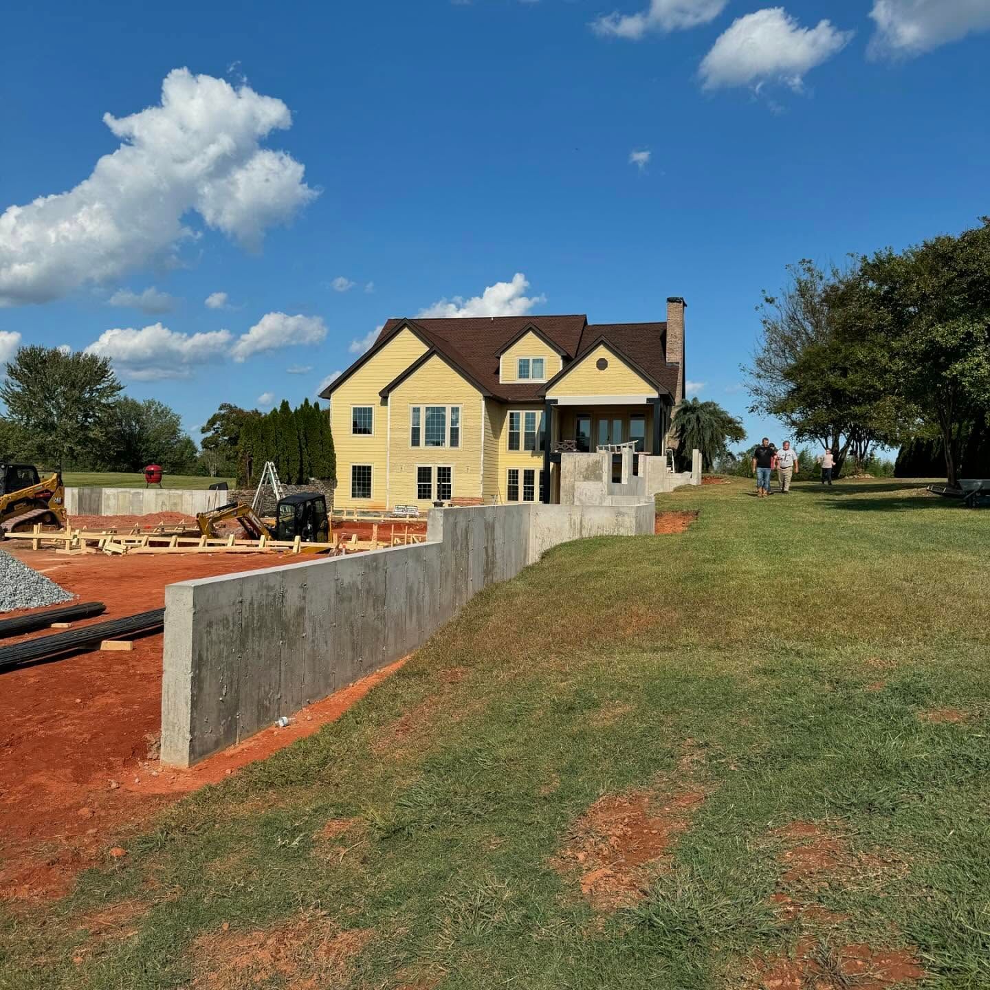 New house under construction behind a concrete wall, with yellow siding, red dirt, and a blue sky