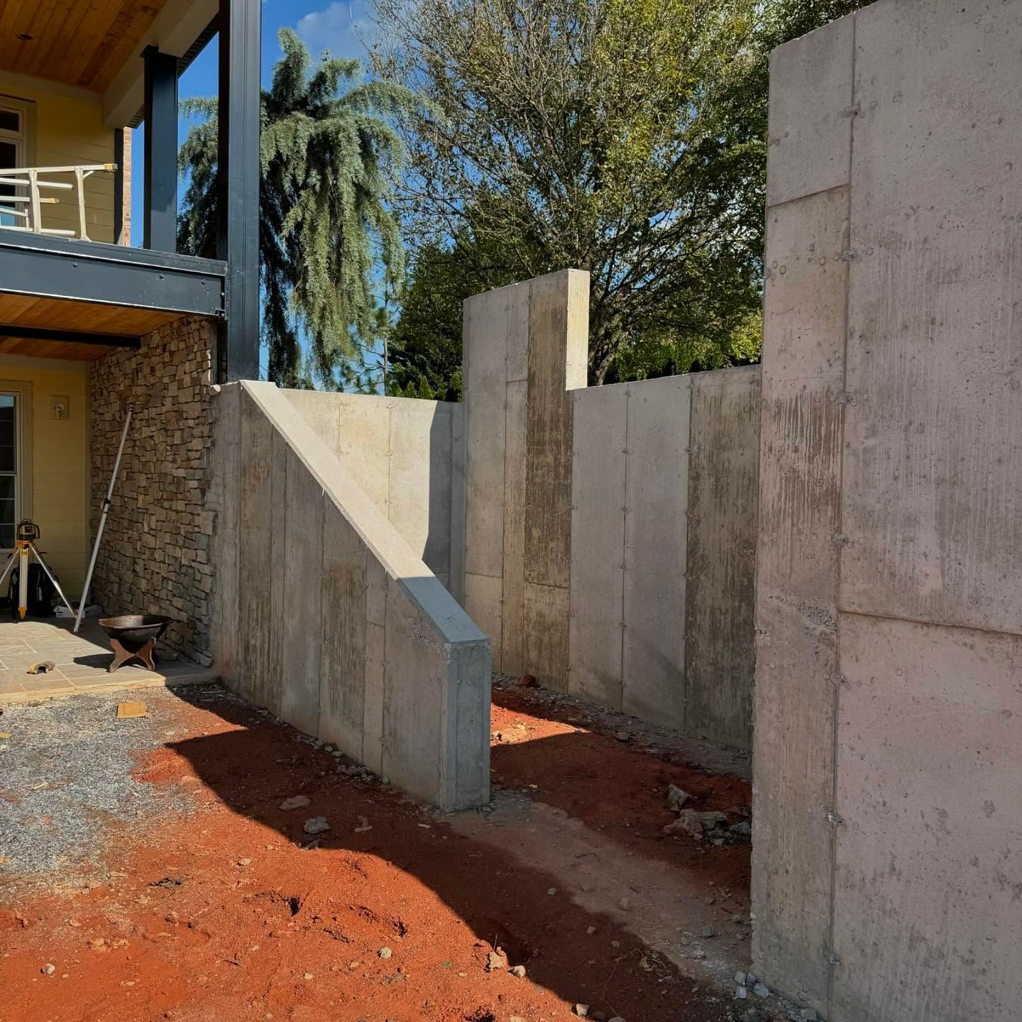 Partially built concrete walls beside a house, with red dirt and trees in the background