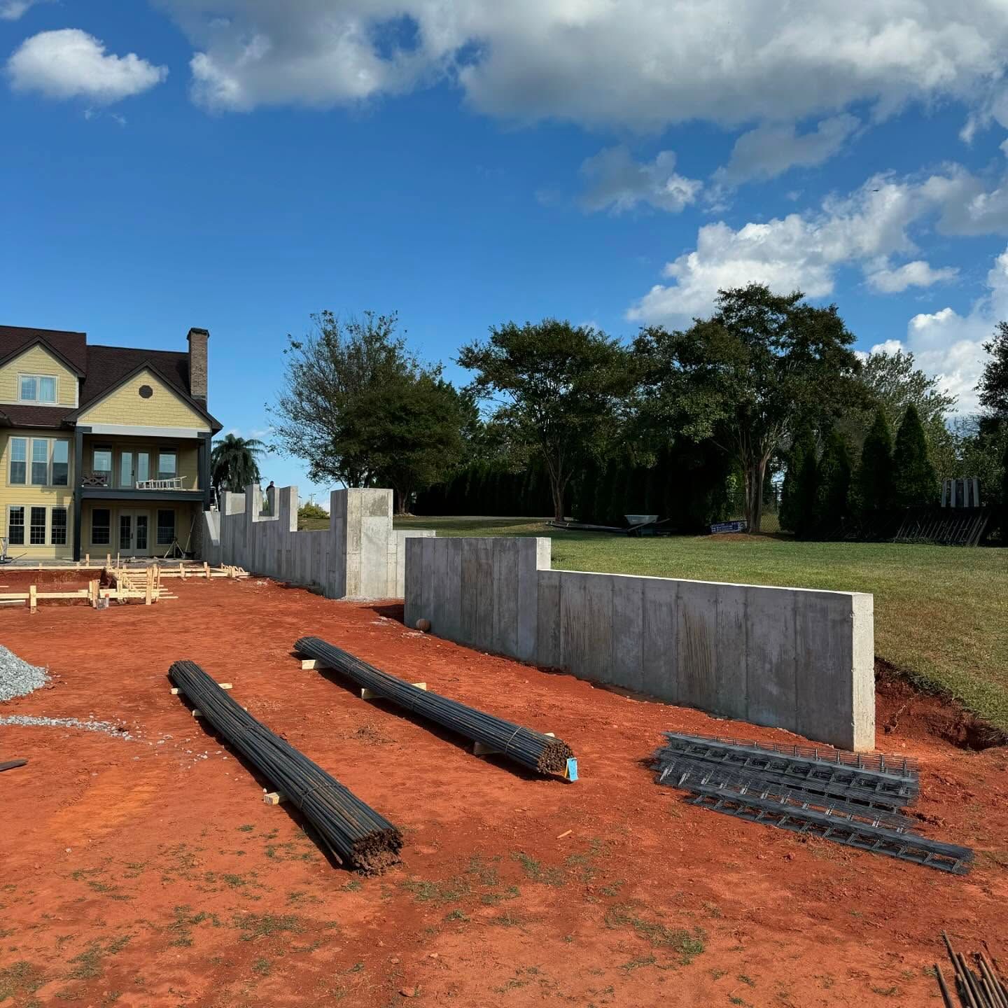 Construction site with stacked black drainage pipes on red dirt beside a house and concrete wall under a blue sky