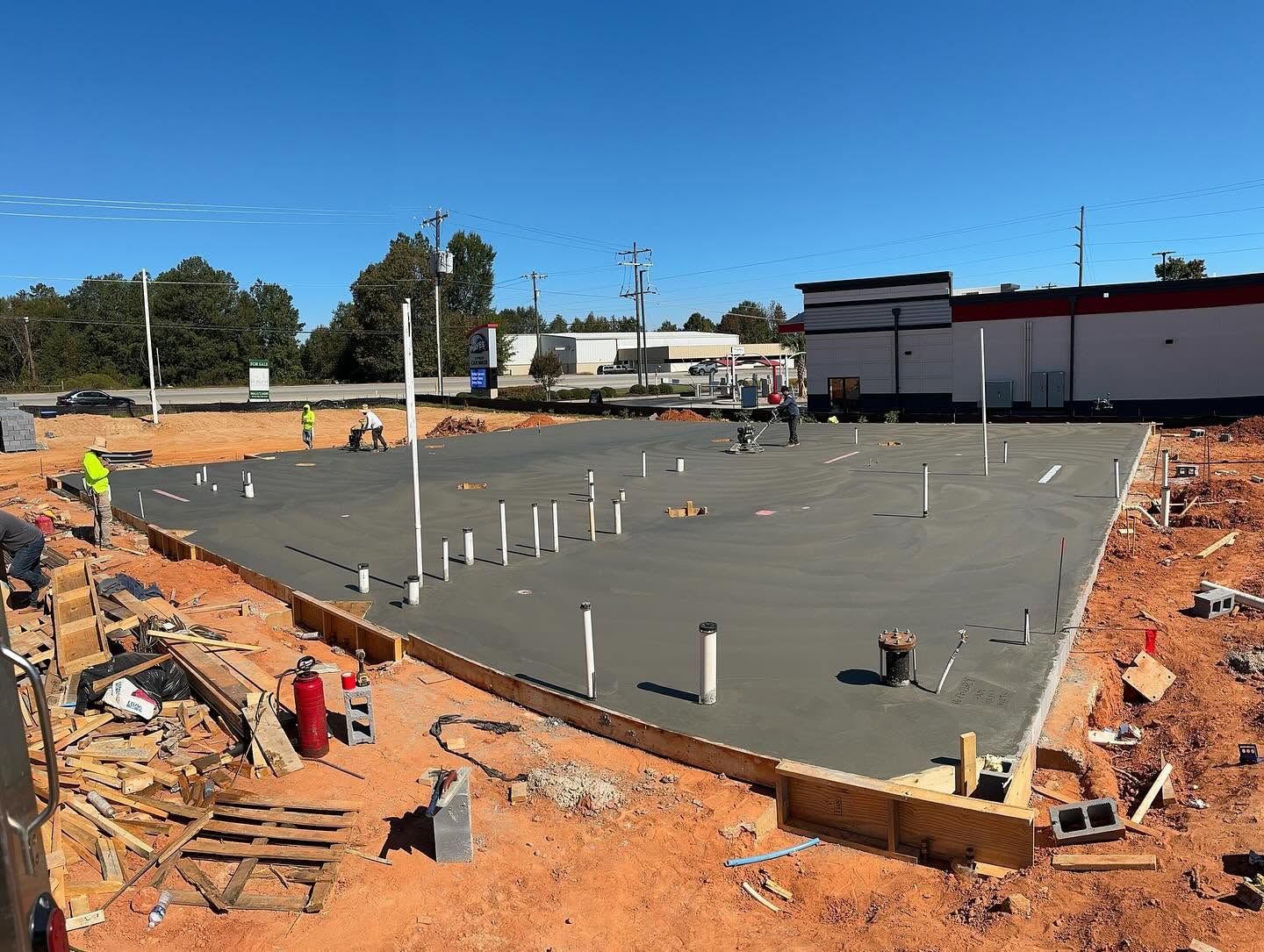 Construction site with fresh concrete foundation, rebar posts, and workers around the perimeter under a clear blue sky