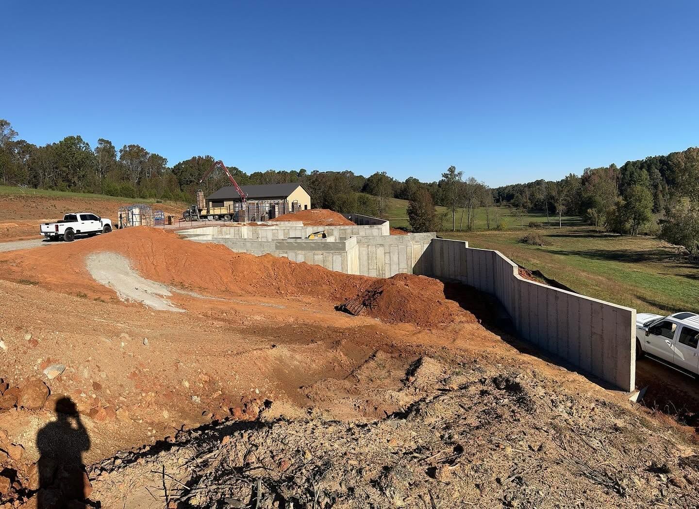 Construction site with excavated red soil, retaining wall, parked trucks, and houses in the background.