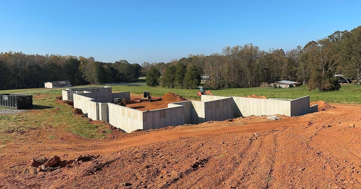 Concrete foundation forms on a dirt lot with a wooded tree line under a clear blue sky