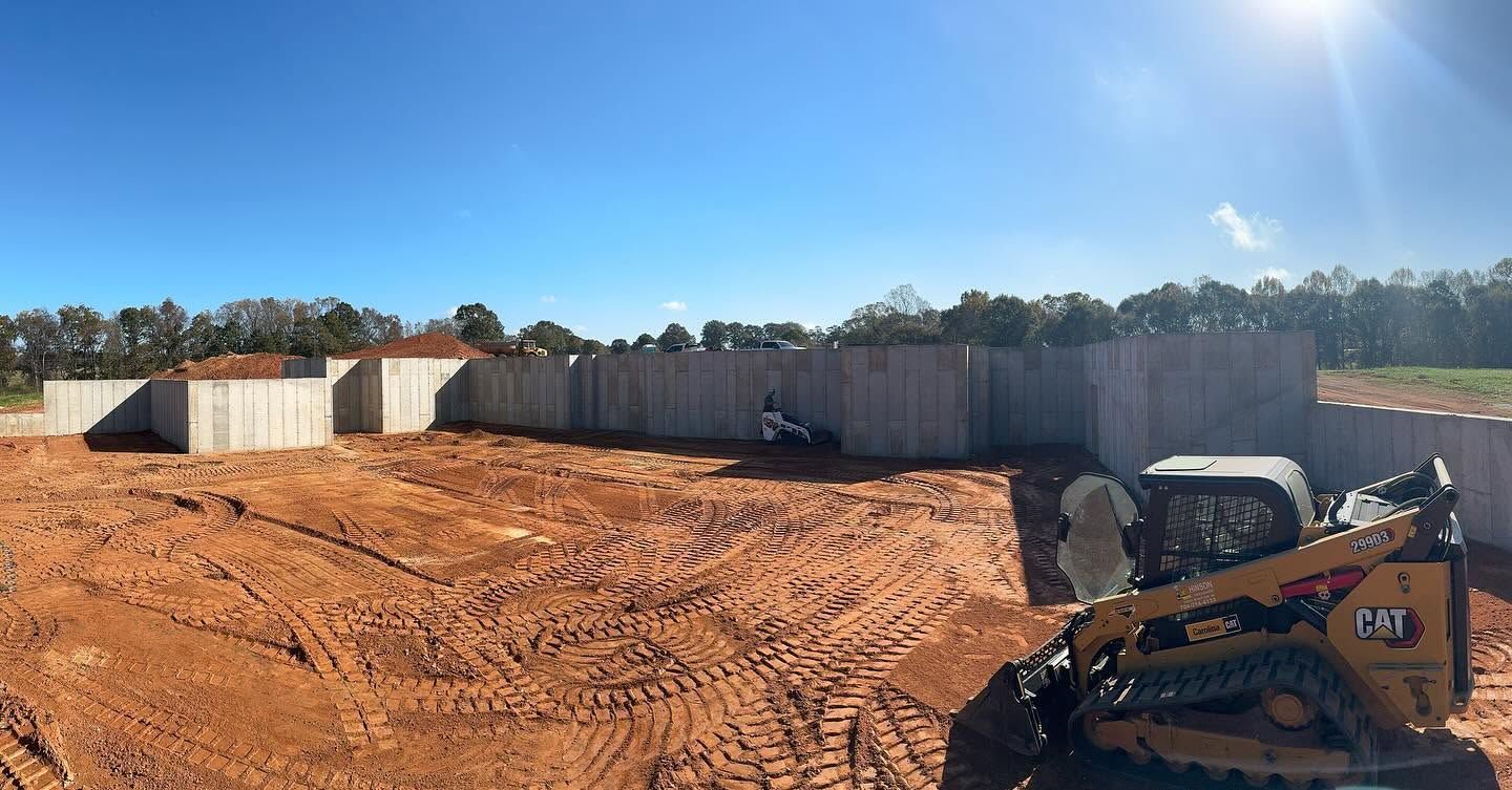 Wide dirt construction site with retaining walls, excavator, and bright sunny sky