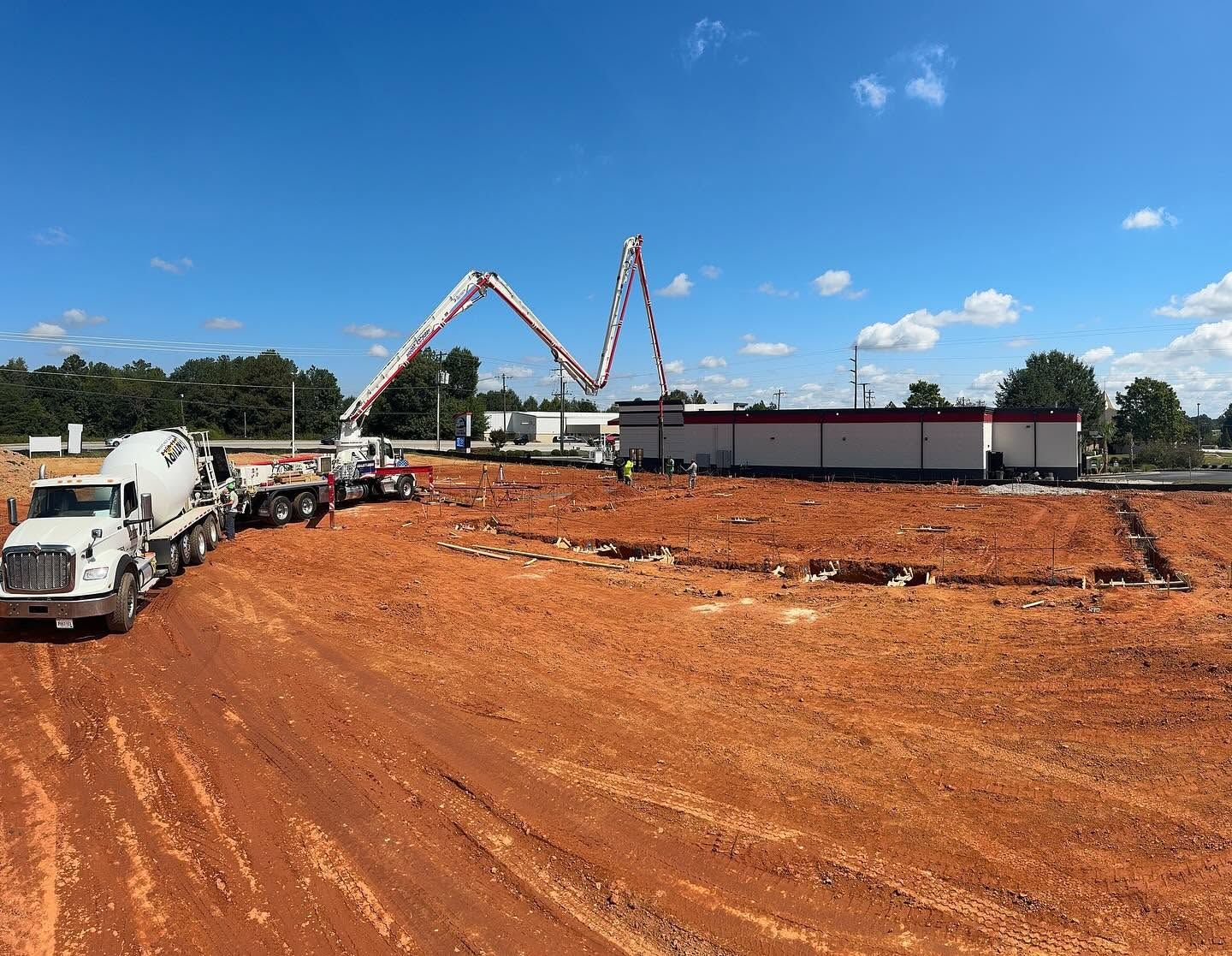 Construction site with trucks and concrete pump booms over a large red dirt lot under a blue sky