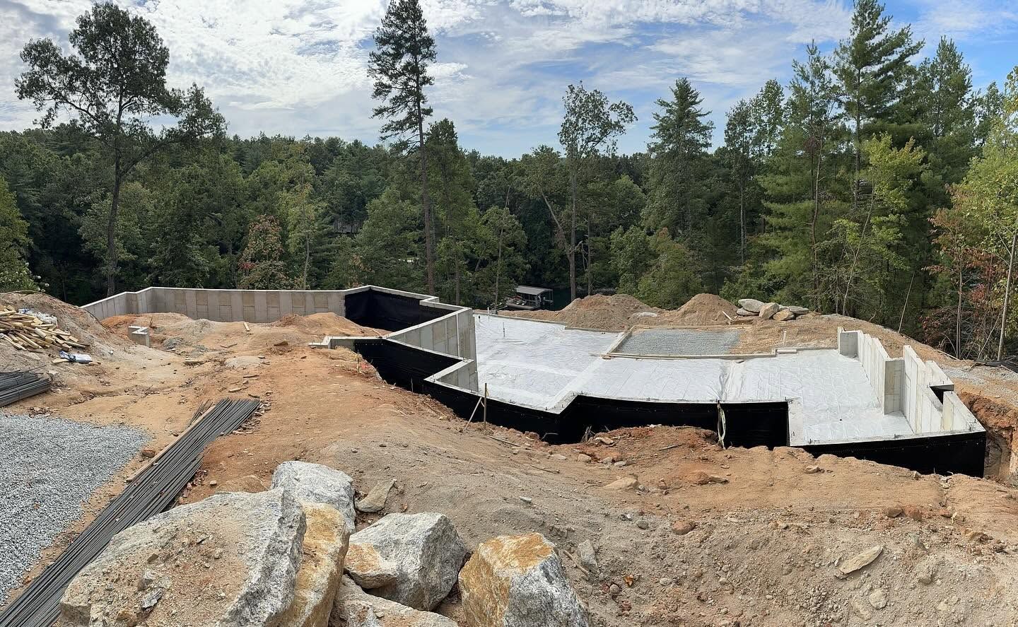 Concrete foundation under construction at a wooded site, with rocks and dirt piles nearby