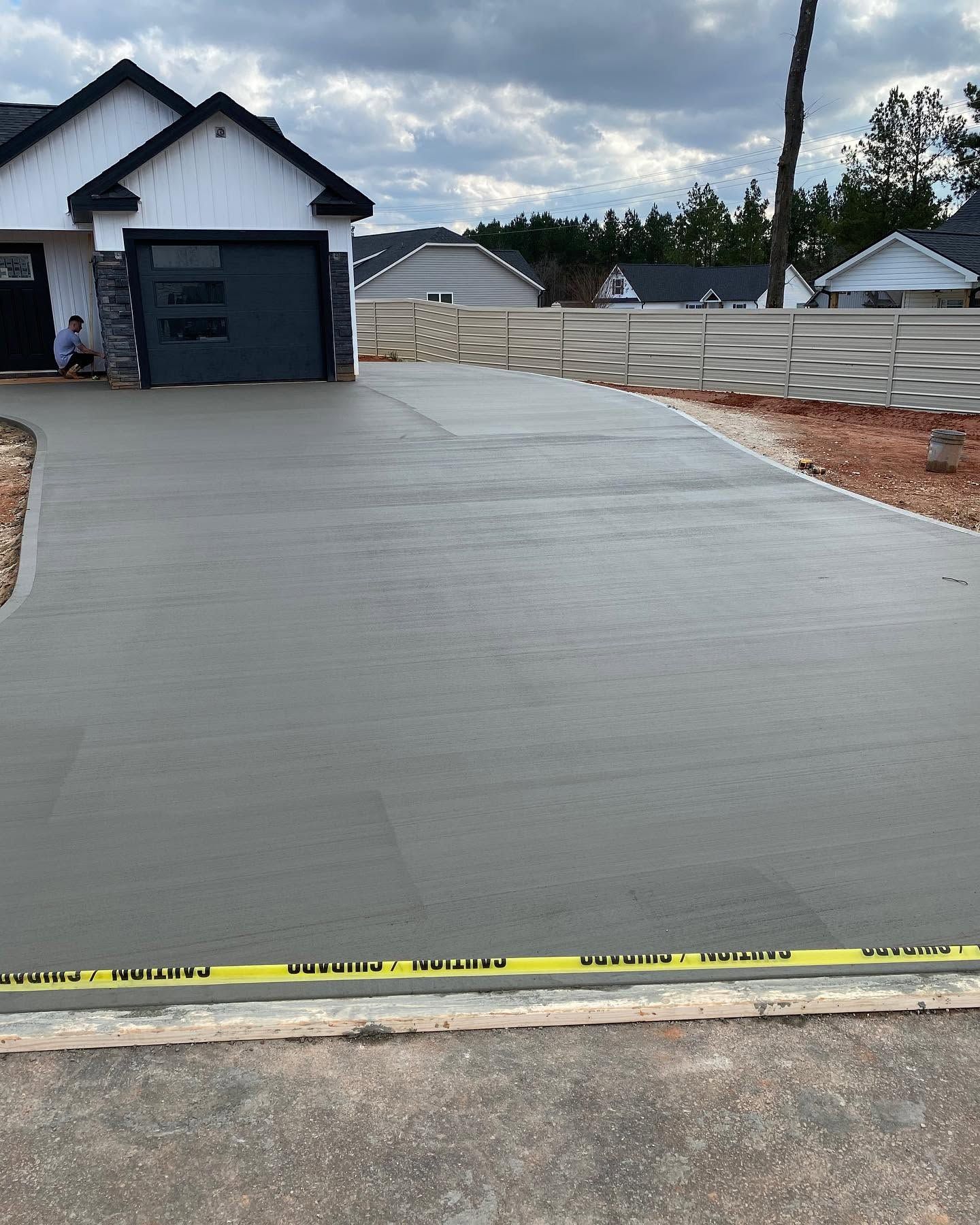 Freshly poured concrete driveway leading to a white house with a black garage door, under construction.
