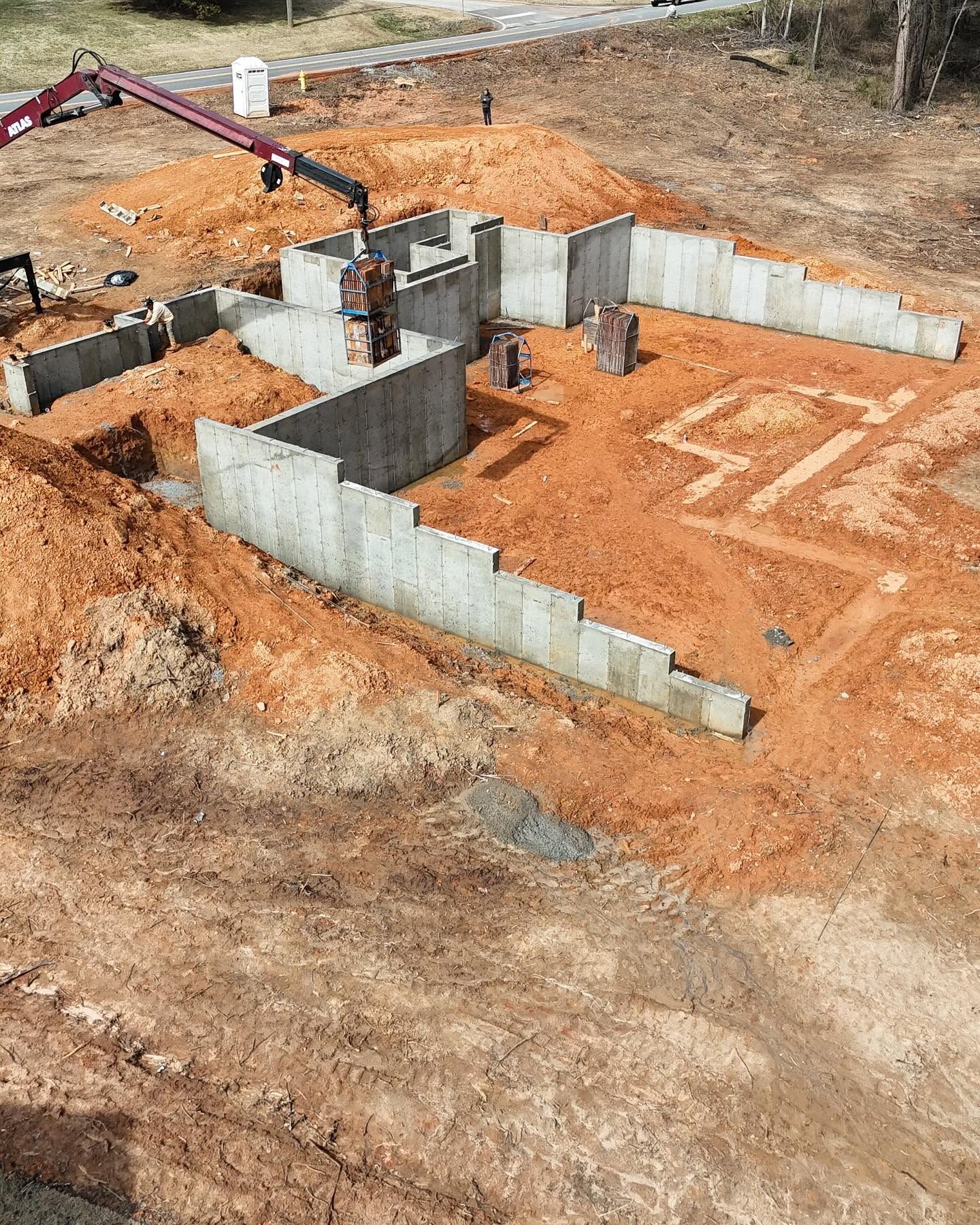 Aerial view of a house foundation under construction on red dirt, with concrete walls and workers present