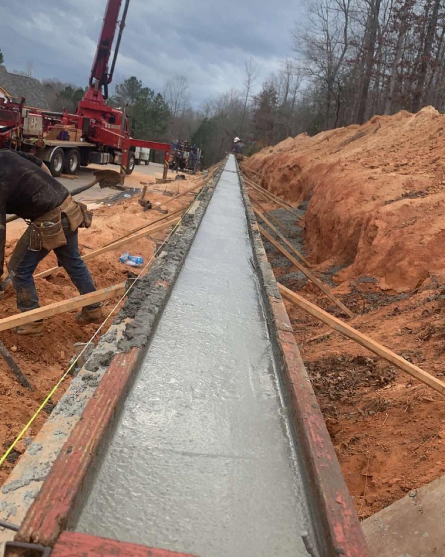 Concrete trench being poured at a construction site, with workers, a pump truck, and dirt embankments.