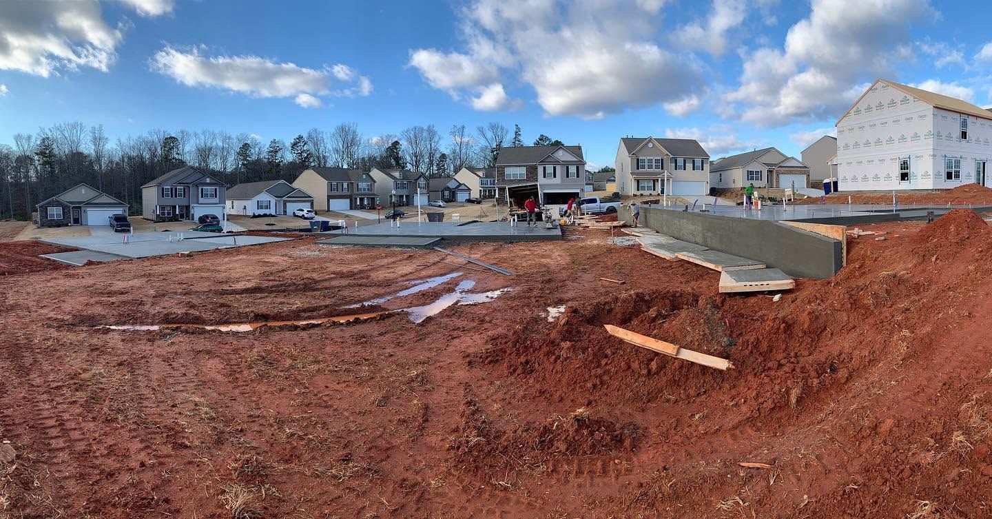 Construction site with red dirt lot, concrete foundation, and houses under a partly cloudy sky