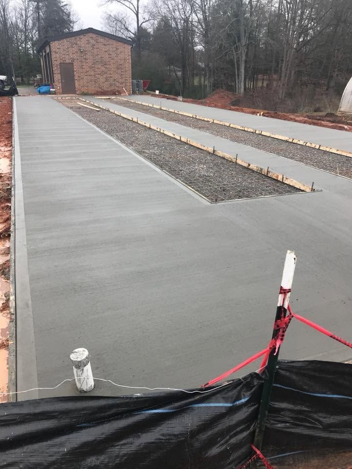 Freshly poured concrete walkway and parking area beside a brick building in a wooded setting