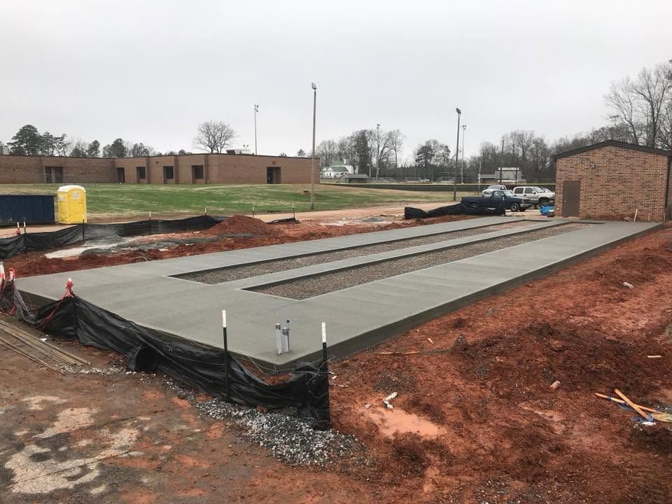Concrete foundation slab under construction beside a brick building, with orange safety fencing and muddy ground