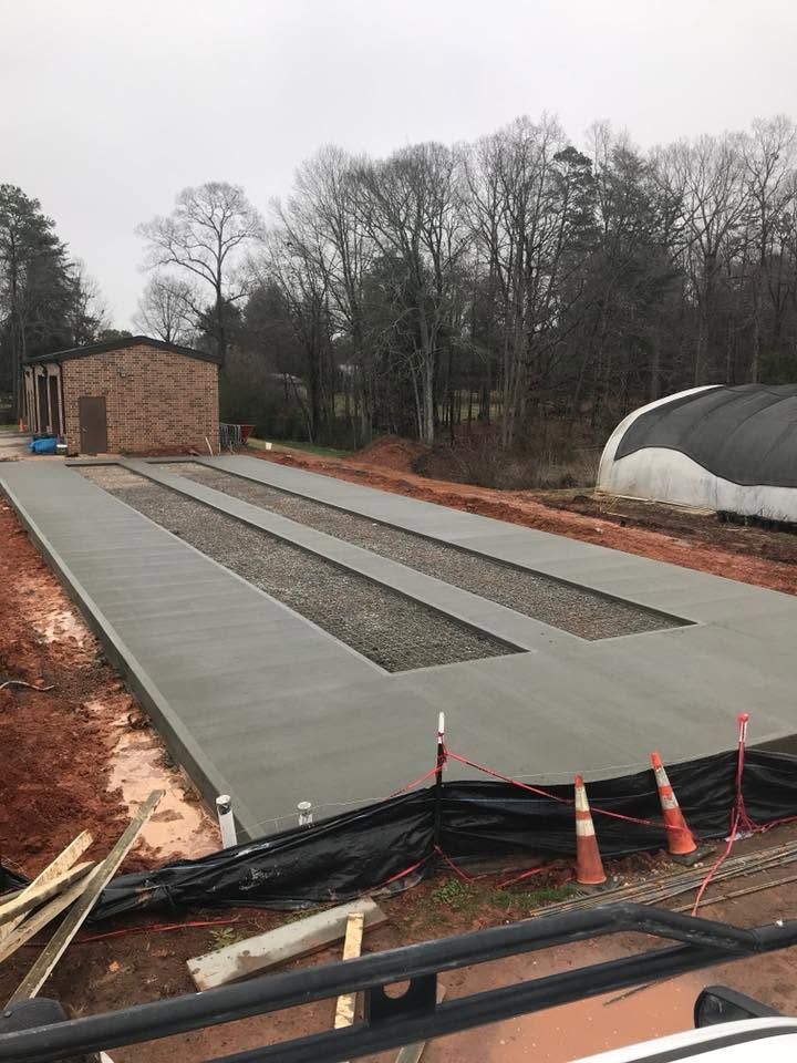 Concrete driveway and narrow gravel strips beside a brick building and wooded area under construction
