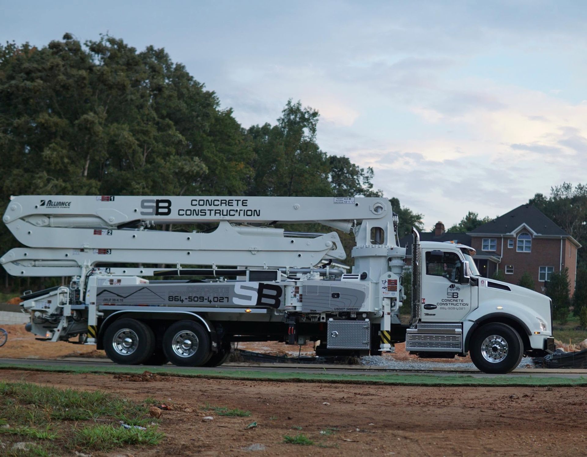 White concrete pump truck on a construction site beside dirt and trees under a cloudy sky