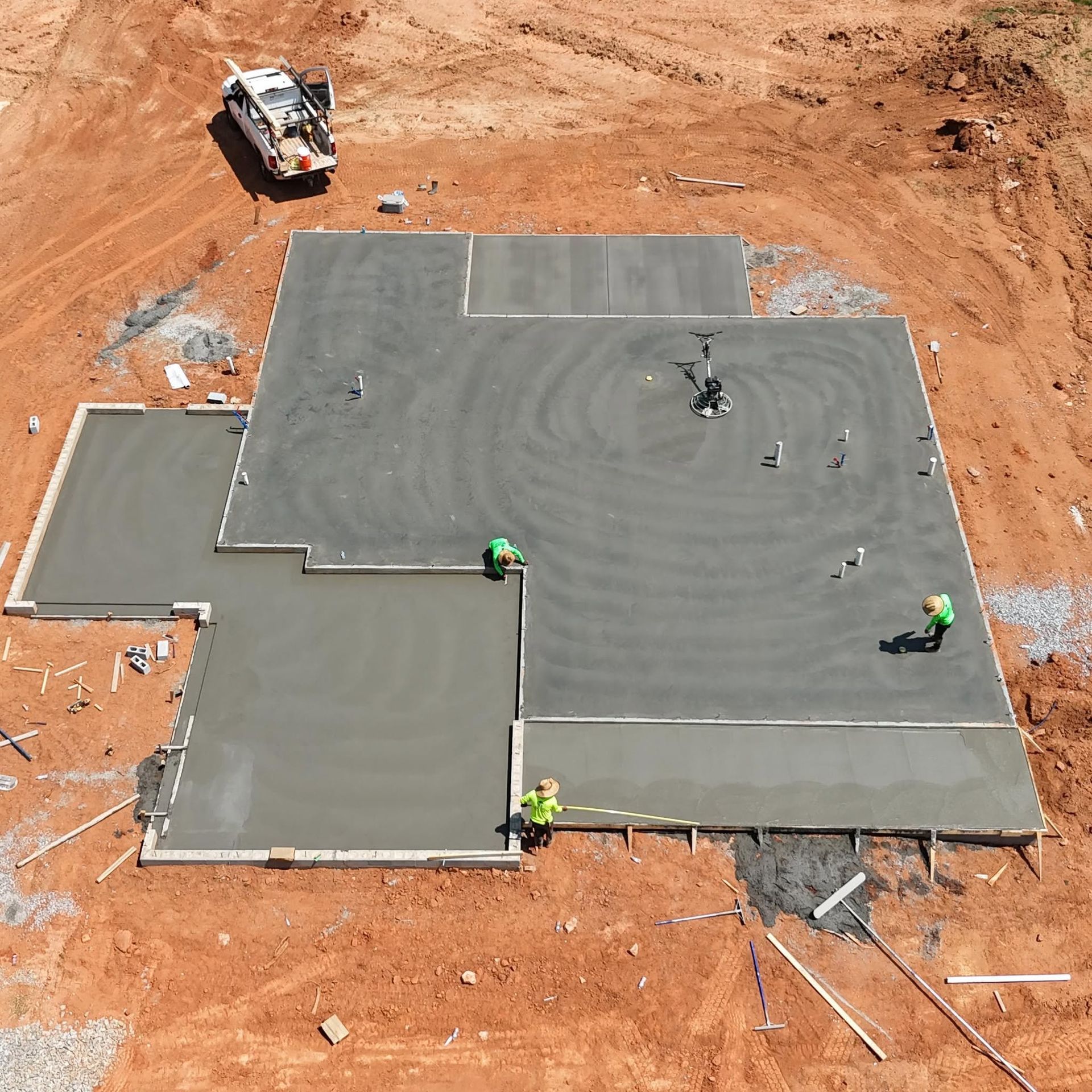 Workers smoothing a large concrete slab at a construction site with a truck and orange dirt around it