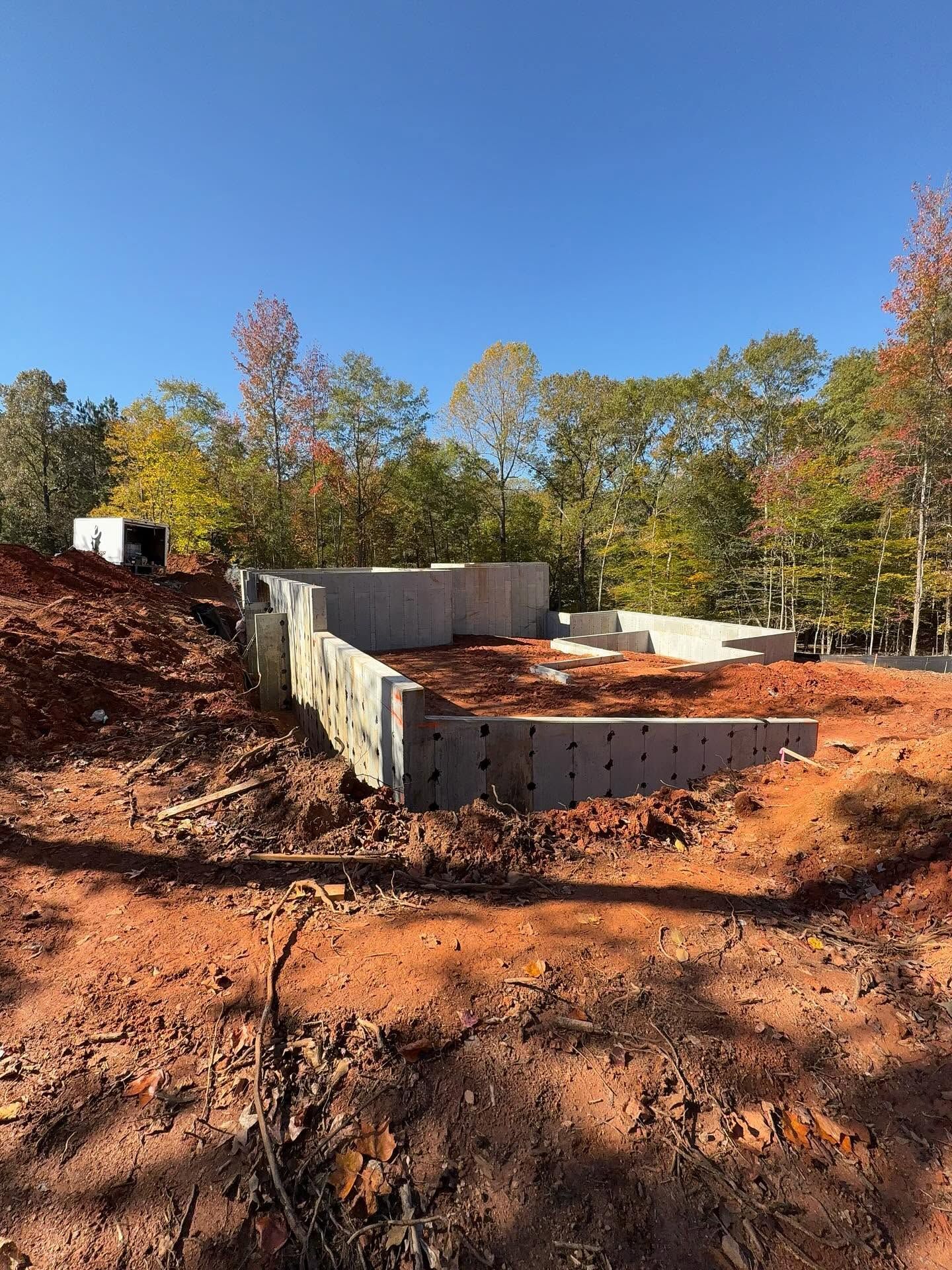 Concrete foundation site in a wooded area with red dirt and autumn trees under a blue sky