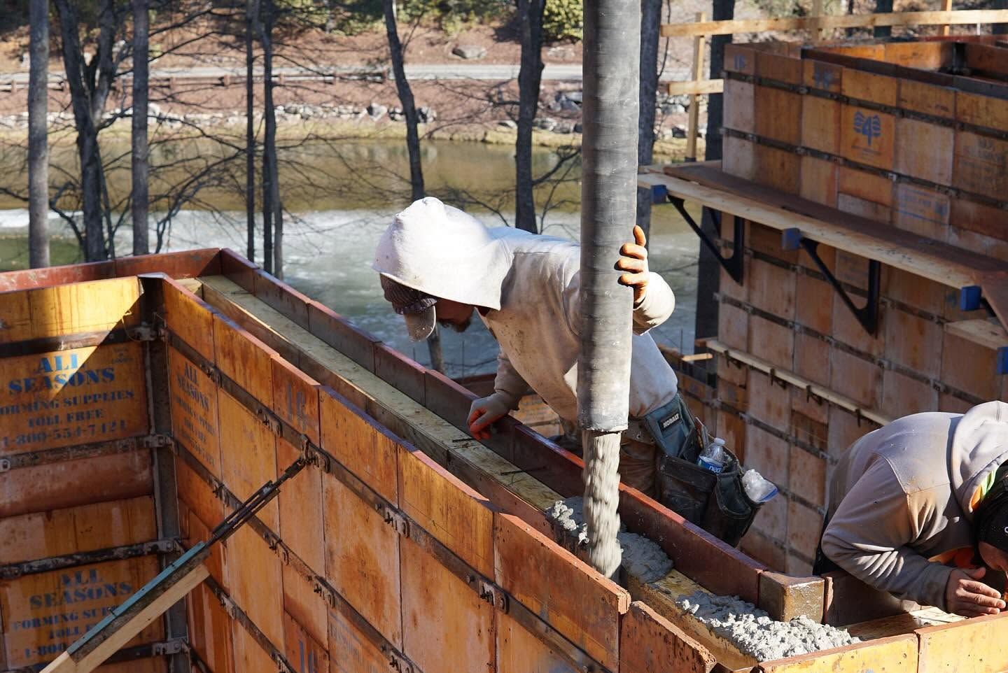 Workers building a concrete foundation beside a wooden form, wearing hard hats at an outdoor construction site