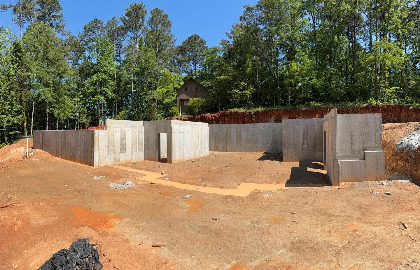 Concrete foundation site on a dirt lot with retaining walls, surrounded by trees and blue sky