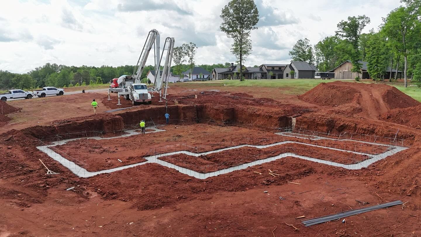 Excavator digging a large foundation in a red dirt construction site, with white layout lines marked on the ground
