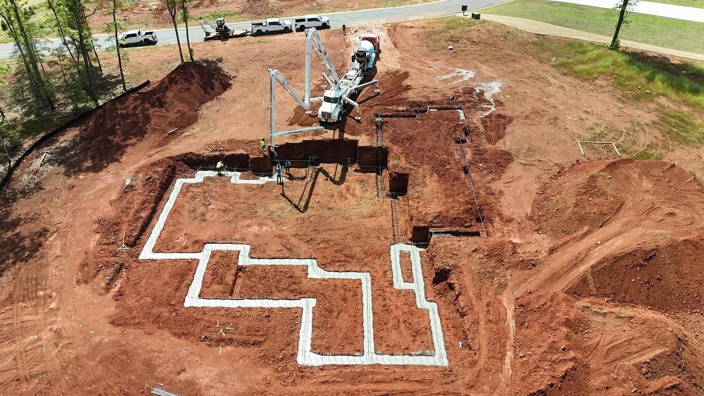 Excavator trenching a red dirt yard with white foundation outlines for a house.
