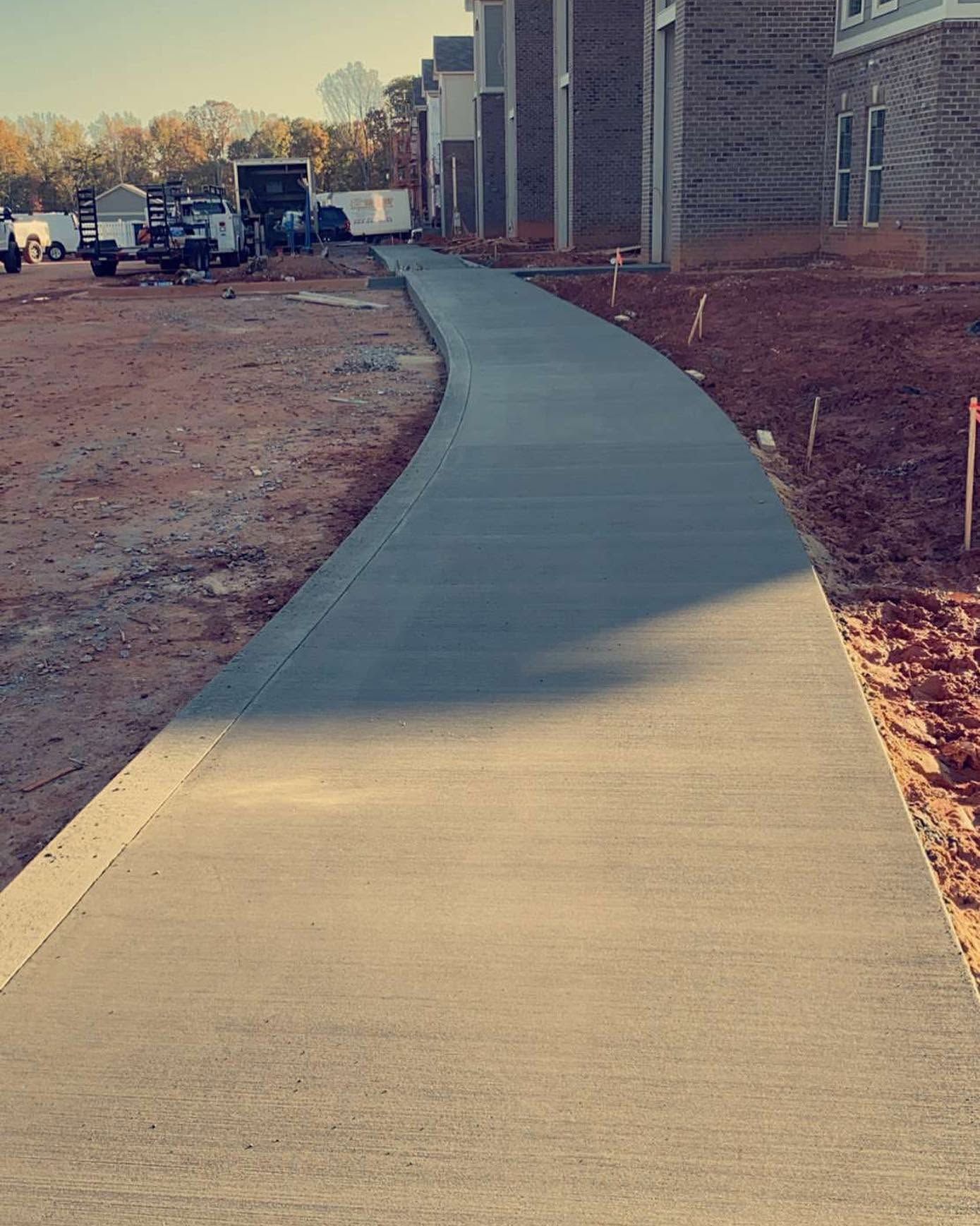 Curved concrete sidewalk beside a building at a construction site, with dirt and equipment ahead.