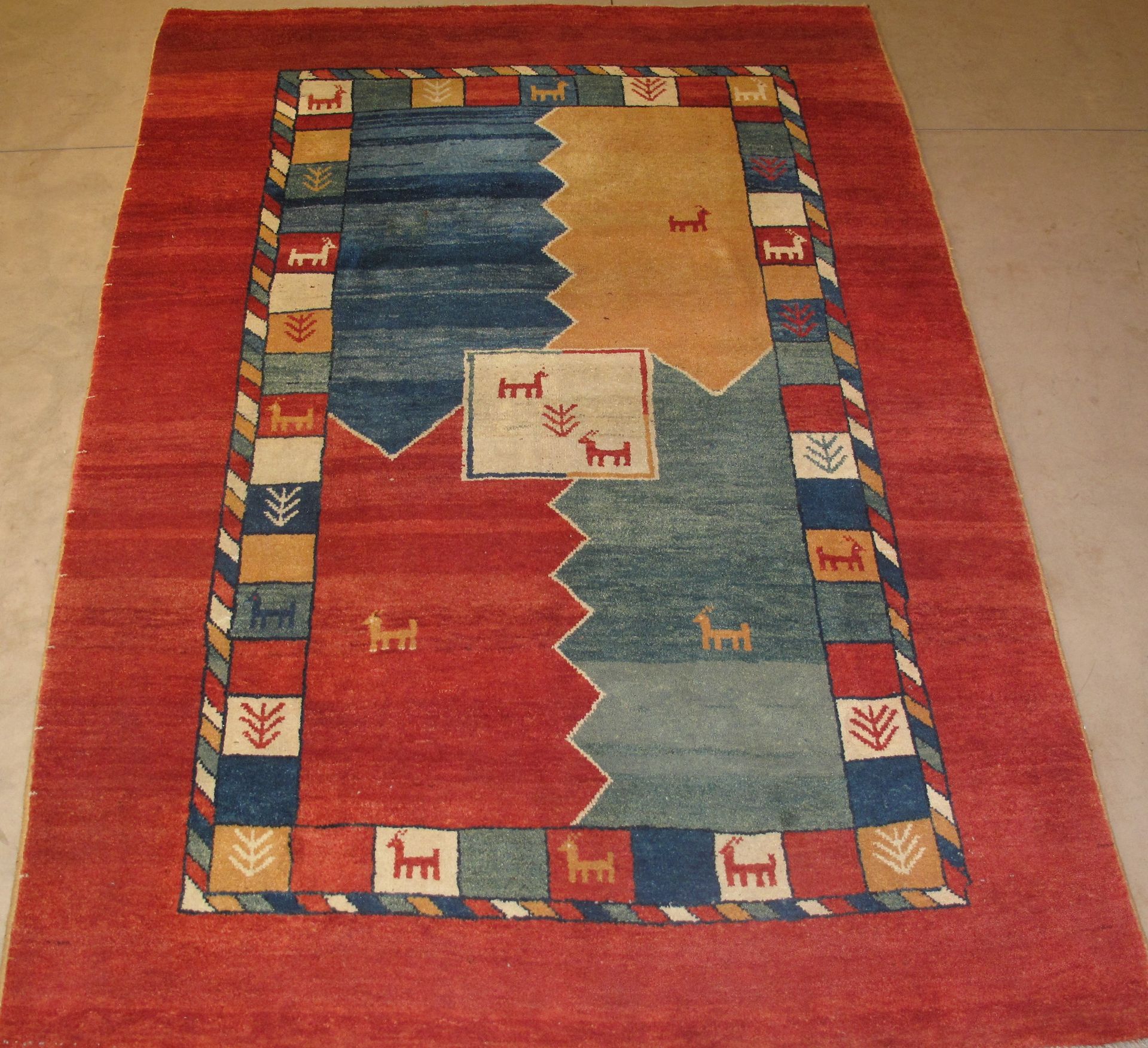 Red rug with blue, tan, and red geometric shapes. Bordered with colorful squares and animal motifs.