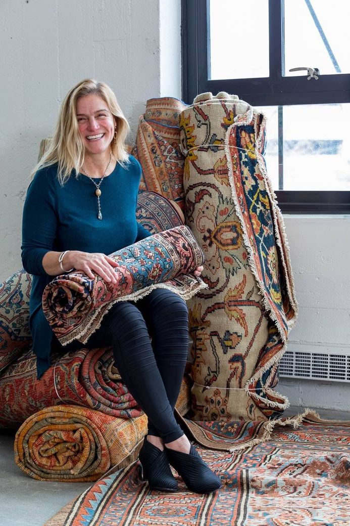 A woman is sitting on a pile of rugs and holding a rug.