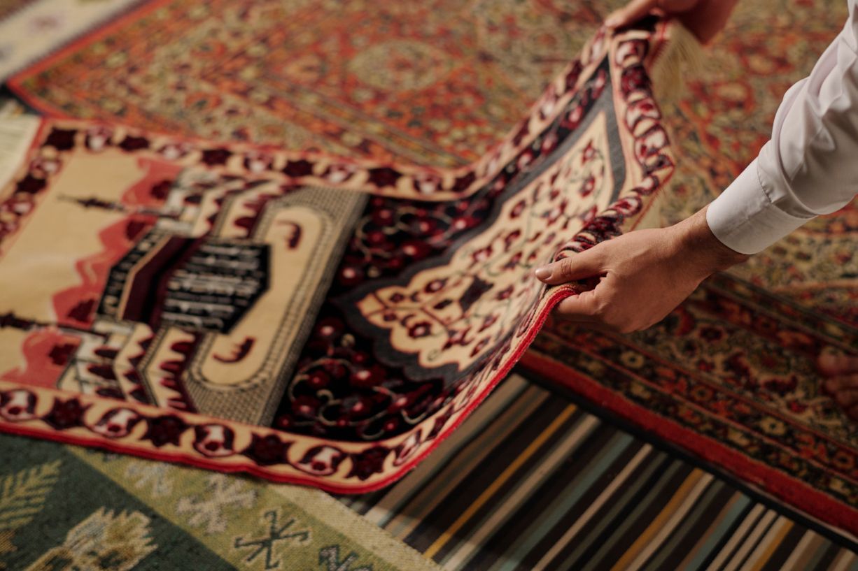 Hands holding an antique rug, unfolding it over other rugs.